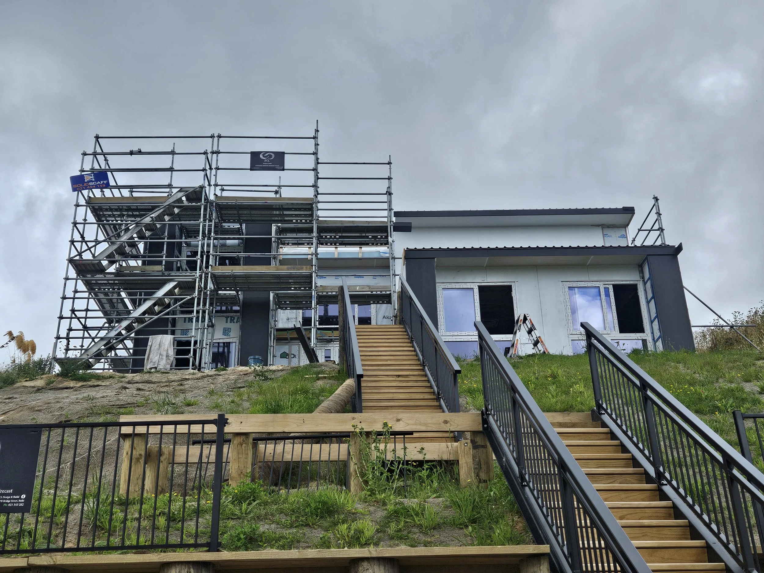 A house under construction with scaffolding around it, a staircase leading to the entrance, a fence in front, and a cloudy sky overhead.
