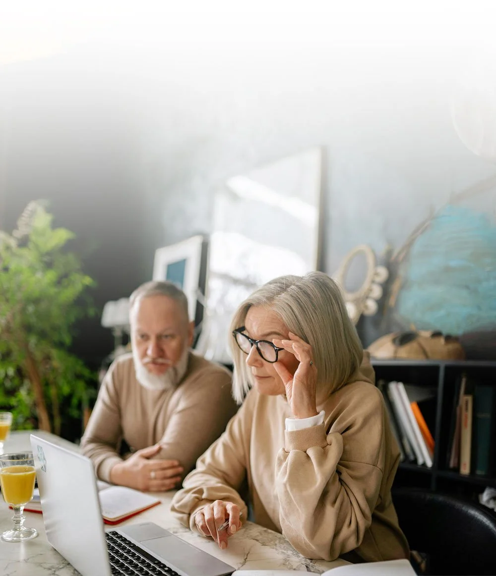 Two older adults, a man and a woman, sitting at a table with a laptop and glasses. The woman is adjusting her glasses and the man is looking at her. There are two glasses of yellow drink on the table, and a notebook. The background is a room with bookshelves and a window with greenery outside.