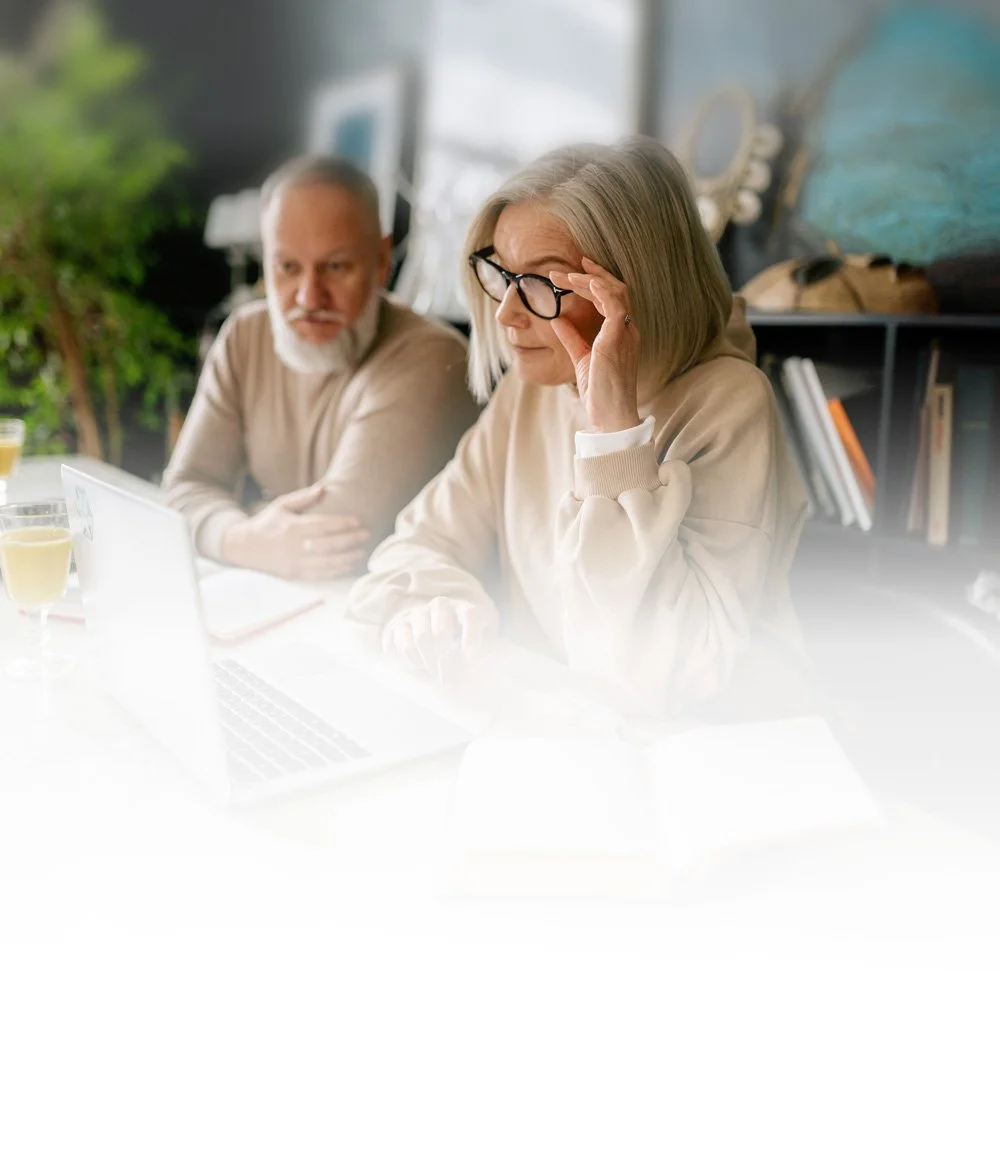 Two older adults, a man and a woman, sitting at a table with a laptop and glasses. The woman is adjusting her glasses and the man is looking at her. There are two glasses of yellow drink on the table, and a notebook. The background is a room with bookshelves and a window with greenery outside.