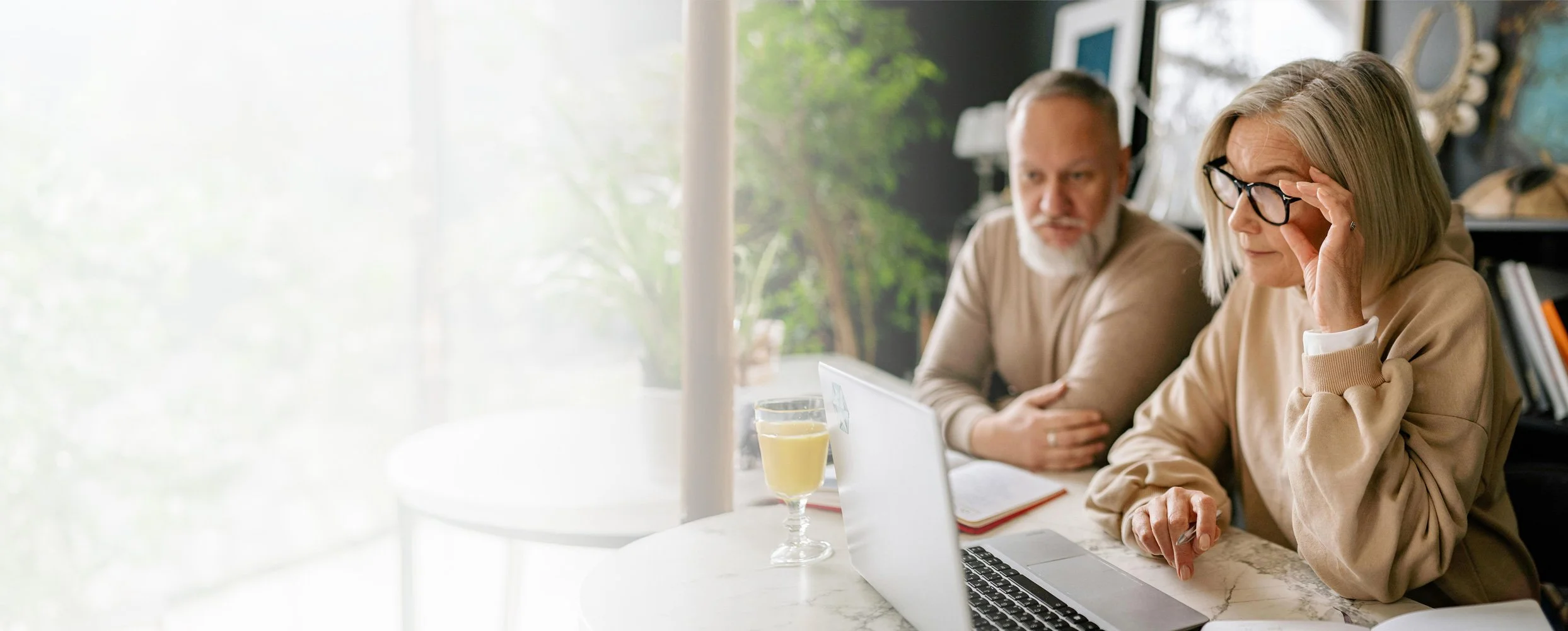 Two older adults, a man and a woman, sitting at a table with a laptop and glasses. The woman is adjusting her glasses and the man is looking at her. There are two glasses of yellow drink on the table, and a notebook. The background is a room with bookshelves and a window with greenery outside.