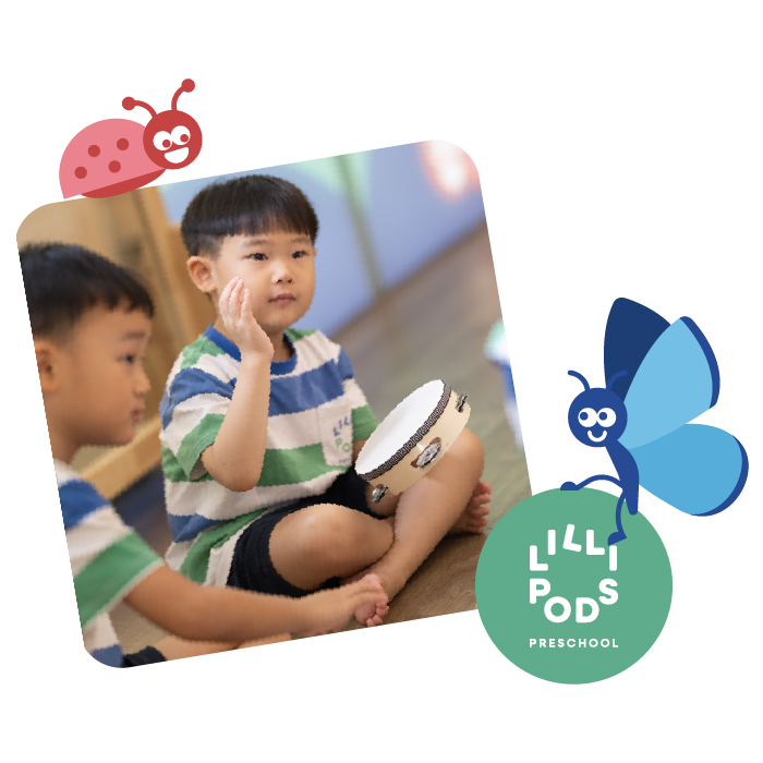 Two young boys sitting on the floor, one is touching his face and the other is reaching for a drum, in a preschool classroom. The image has colorful playful design elements, including a ladybug and a blue butterfly, with the words 'LILIPODS PRESCHOOL' in the bottom right corner.