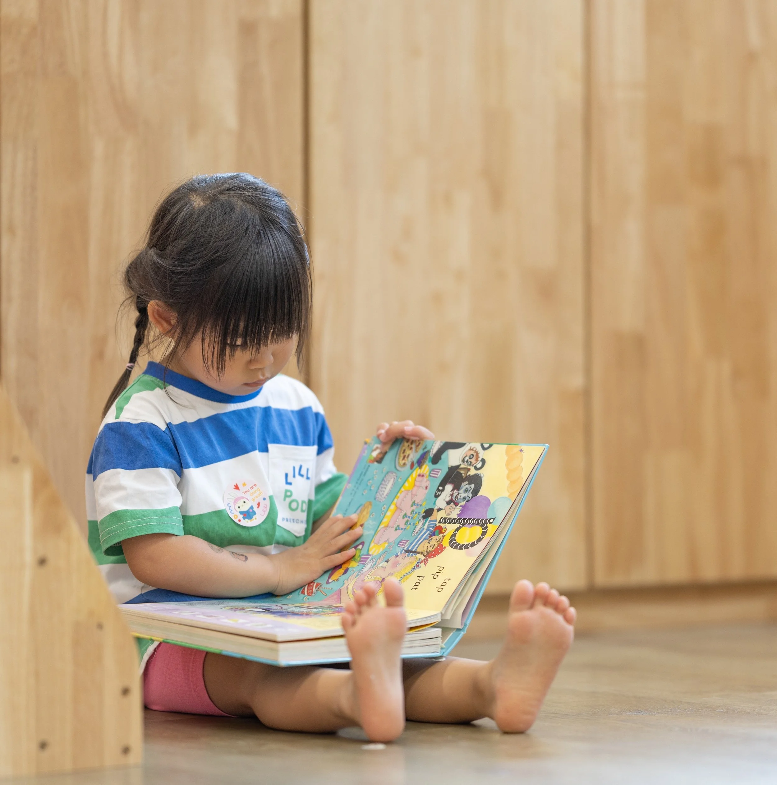 A young girl with black hair, wearing a striped t-shirt, sitting cross-legged on the floor, reading a colorful children's book.