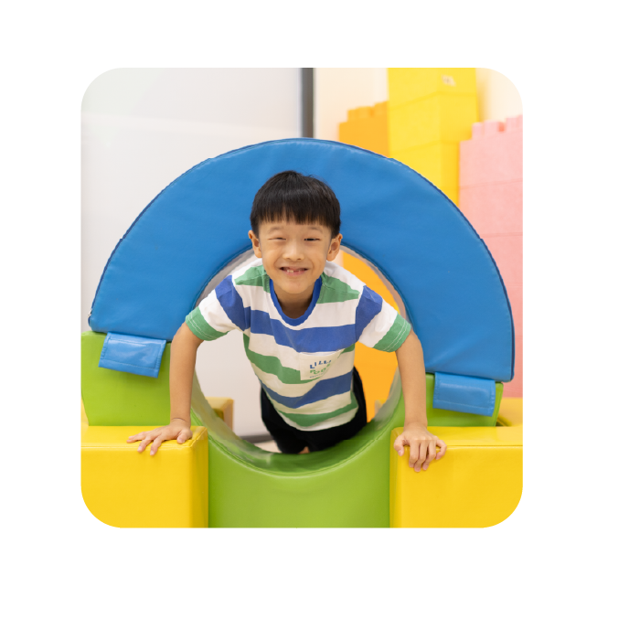 Child smiling while playing in an indoor soft play area with colorful foam blocks and a blue padded tunnel.