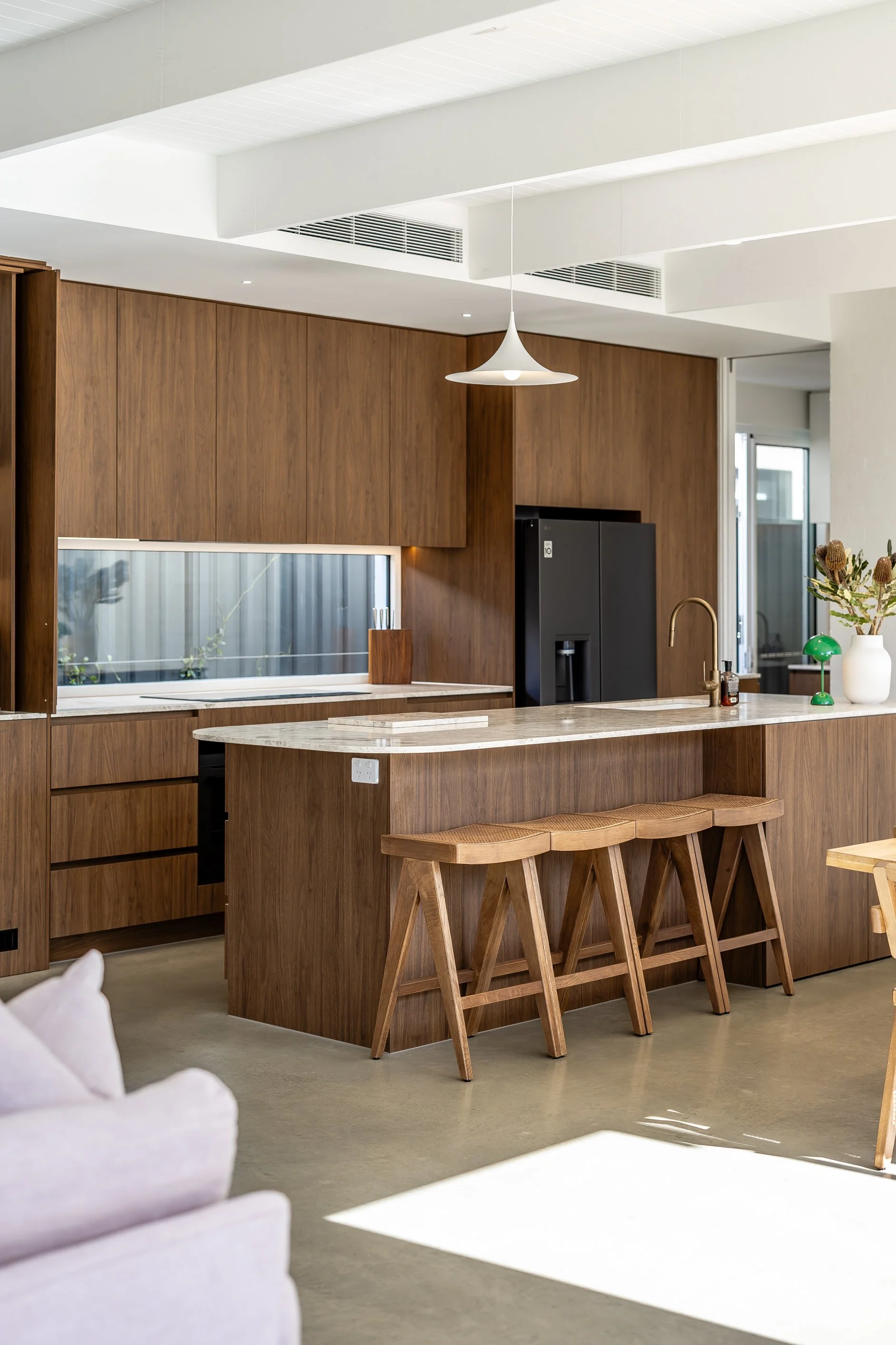 Modern kitchen with wooden cabinets, a marble island with four wooden stools, a brass faucet, and a black refrigerator. Decor includes a green desk lamp and a white vase with flowers. Bright natural light streams in through windows.
