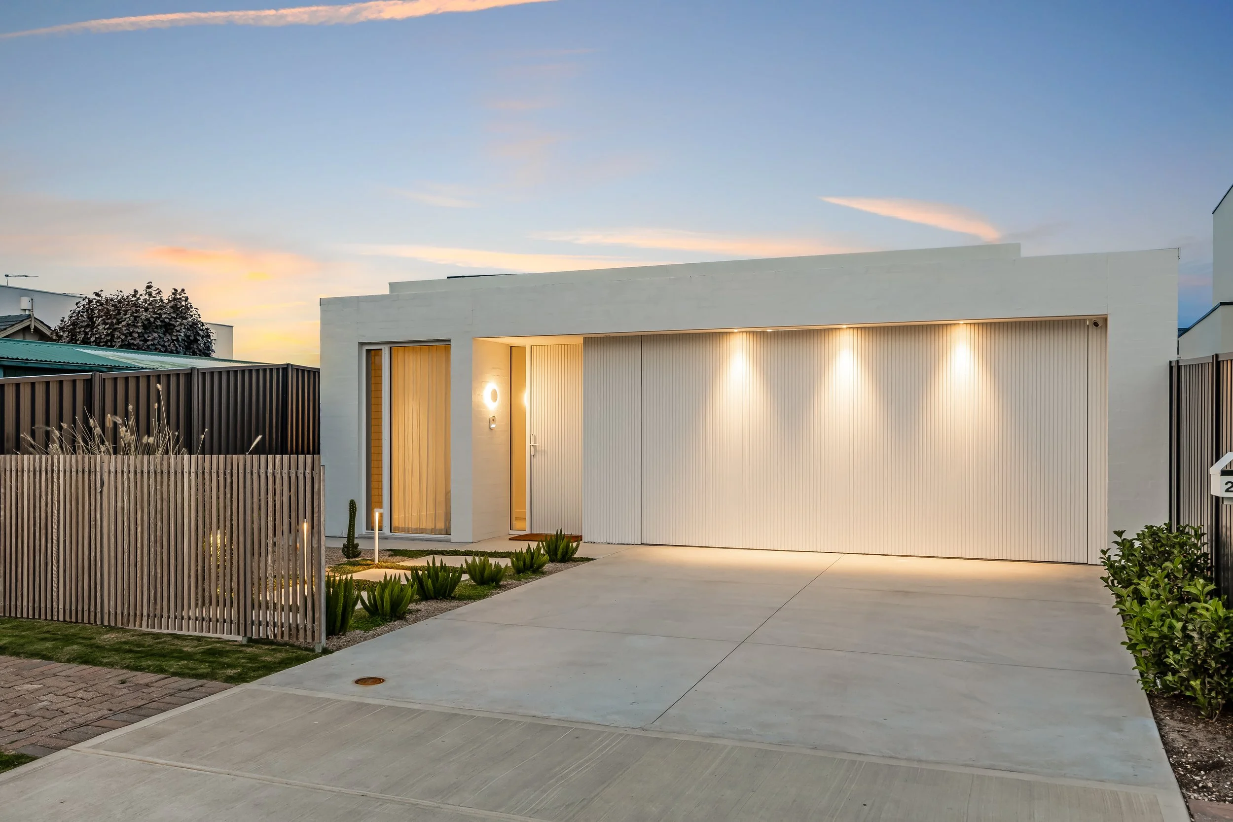 Modern house during sunset with a white facade, wooden accents, and landscaping including small plants and trees, surrounded by a concrete driveway and fences.
