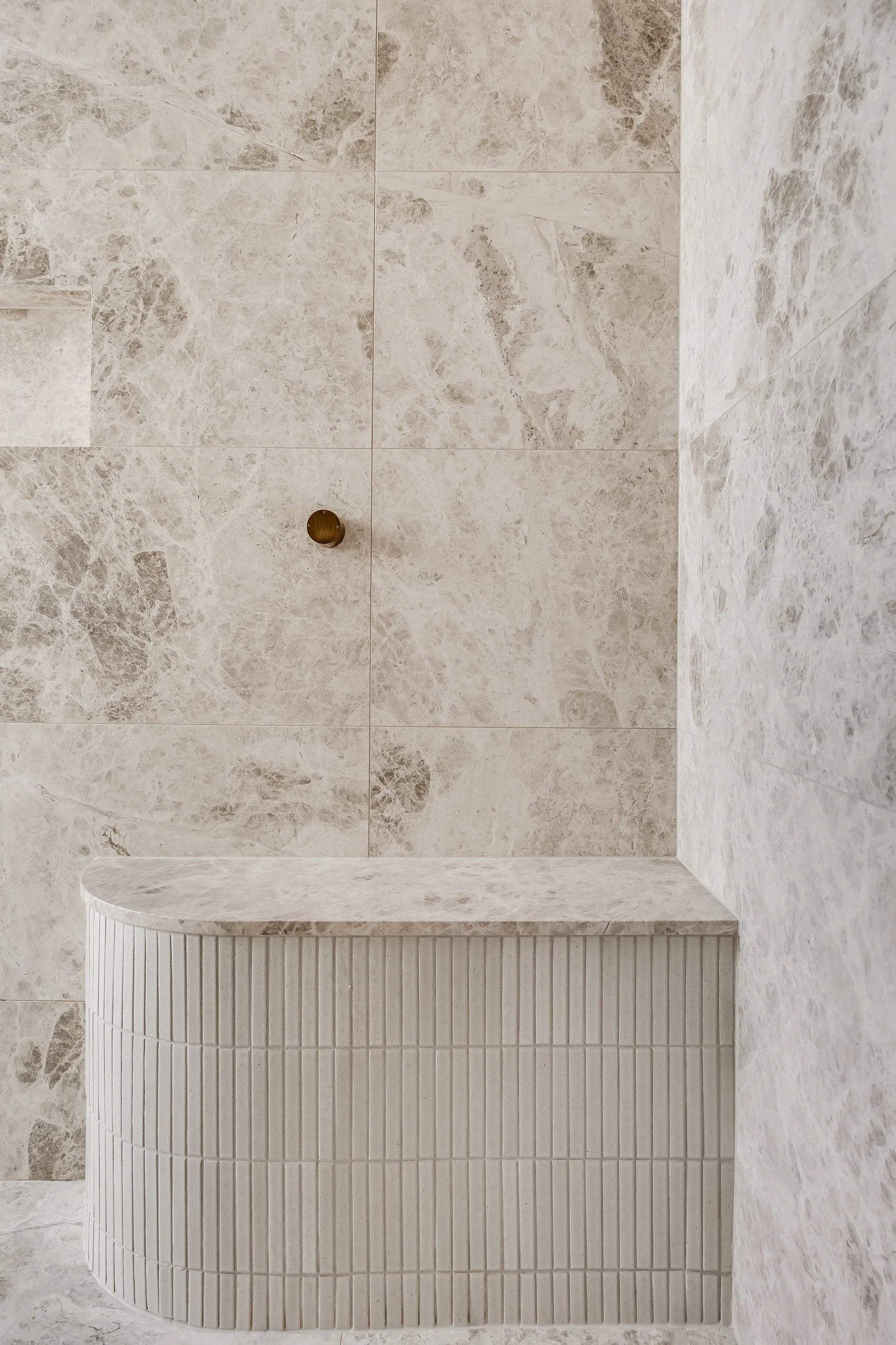 A shower area with marble tiles on the walls and floor, featuring a brass shower drain and a built-in marble seating area with vertical textured ridges on the front.
