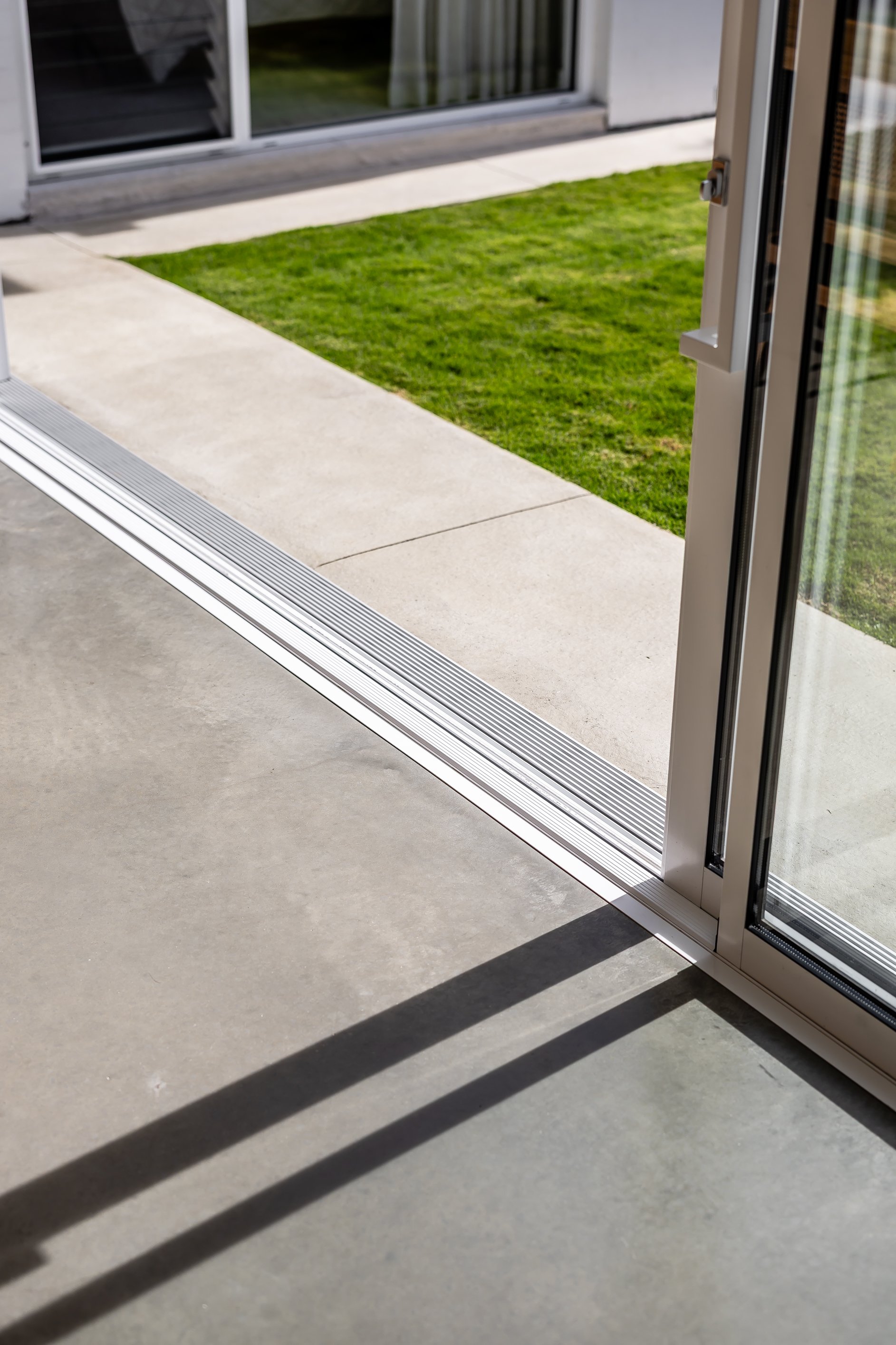 Open sliding glass door leading to a concrete patio and a small patch of green grass outside. Shadows of the door frame are cast on the patio.