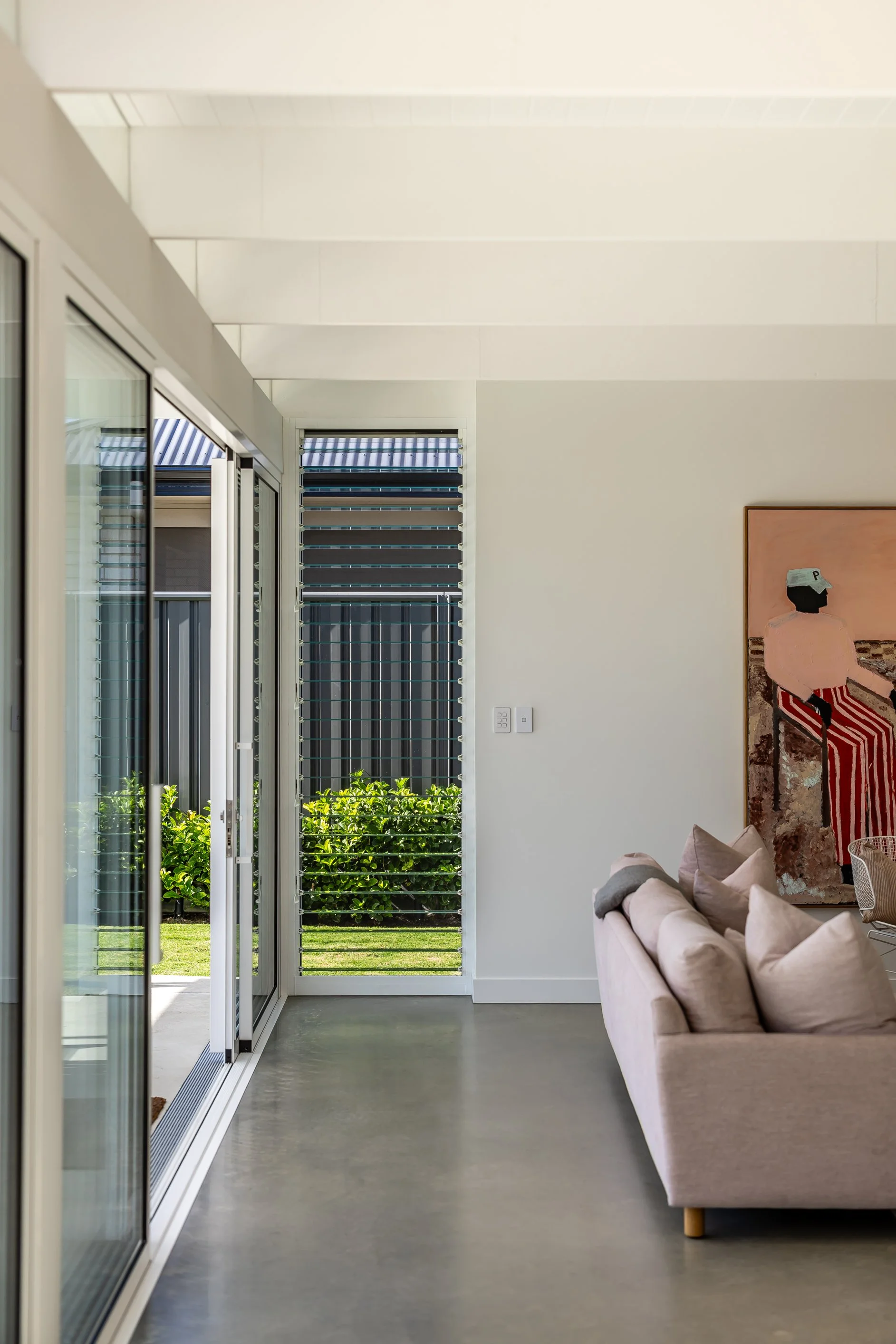 Interior view of a modern living room with a beige sofa, artwork on the wall, and large glass sliding doors opening to a garden with green plants and a lawn.