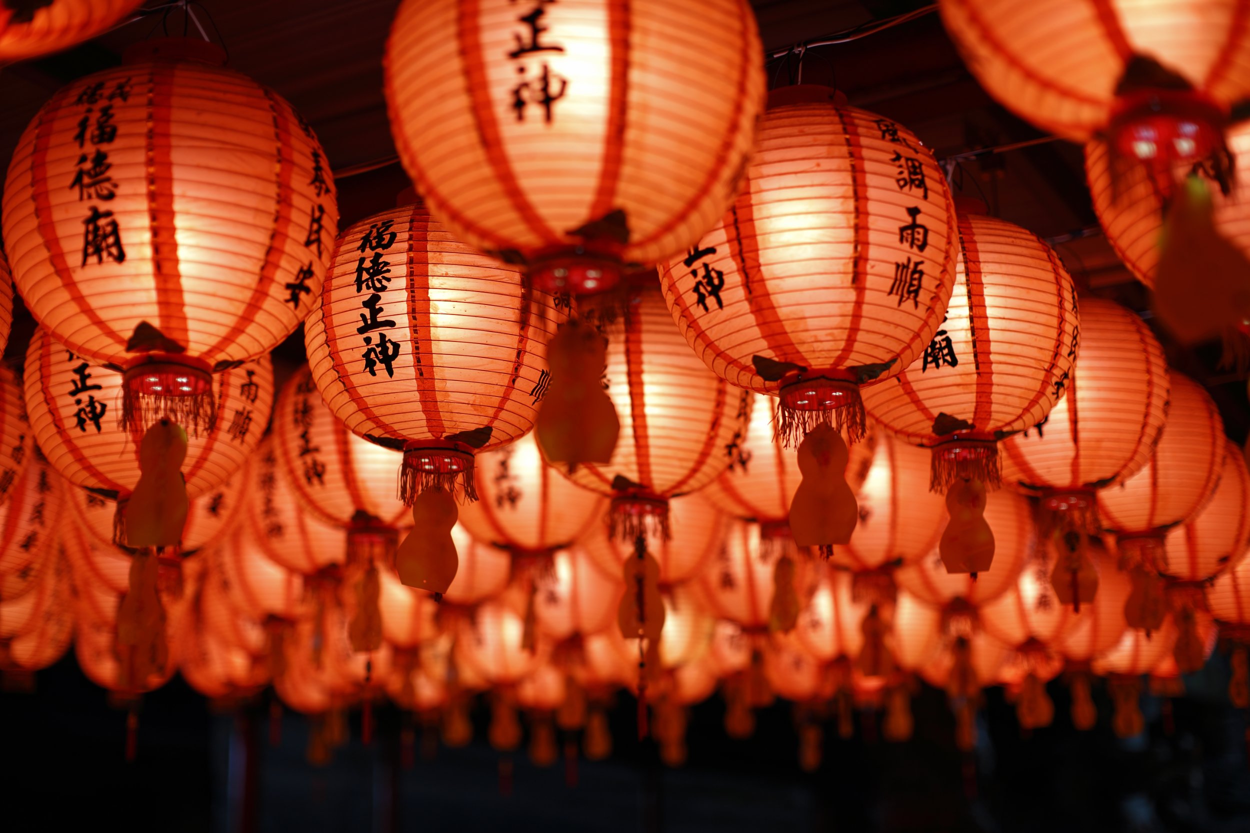Multiple red paper lanterns with black Chinese characters hanging in a row, glowing in warm light.