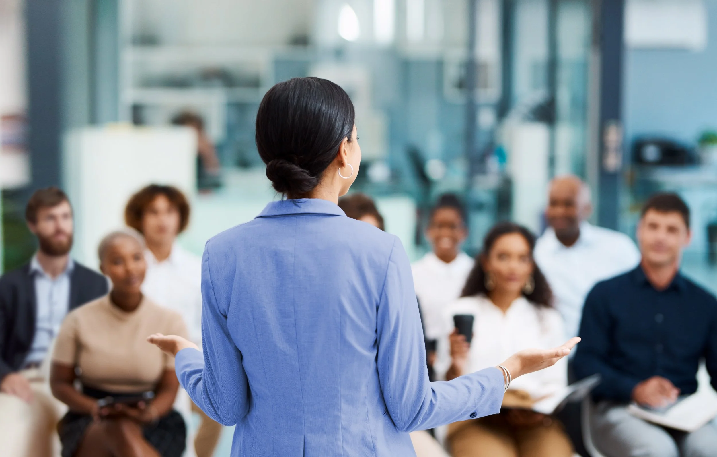 A woman in a blue blazer giving a presentation to a group of diverse people in a modern office.