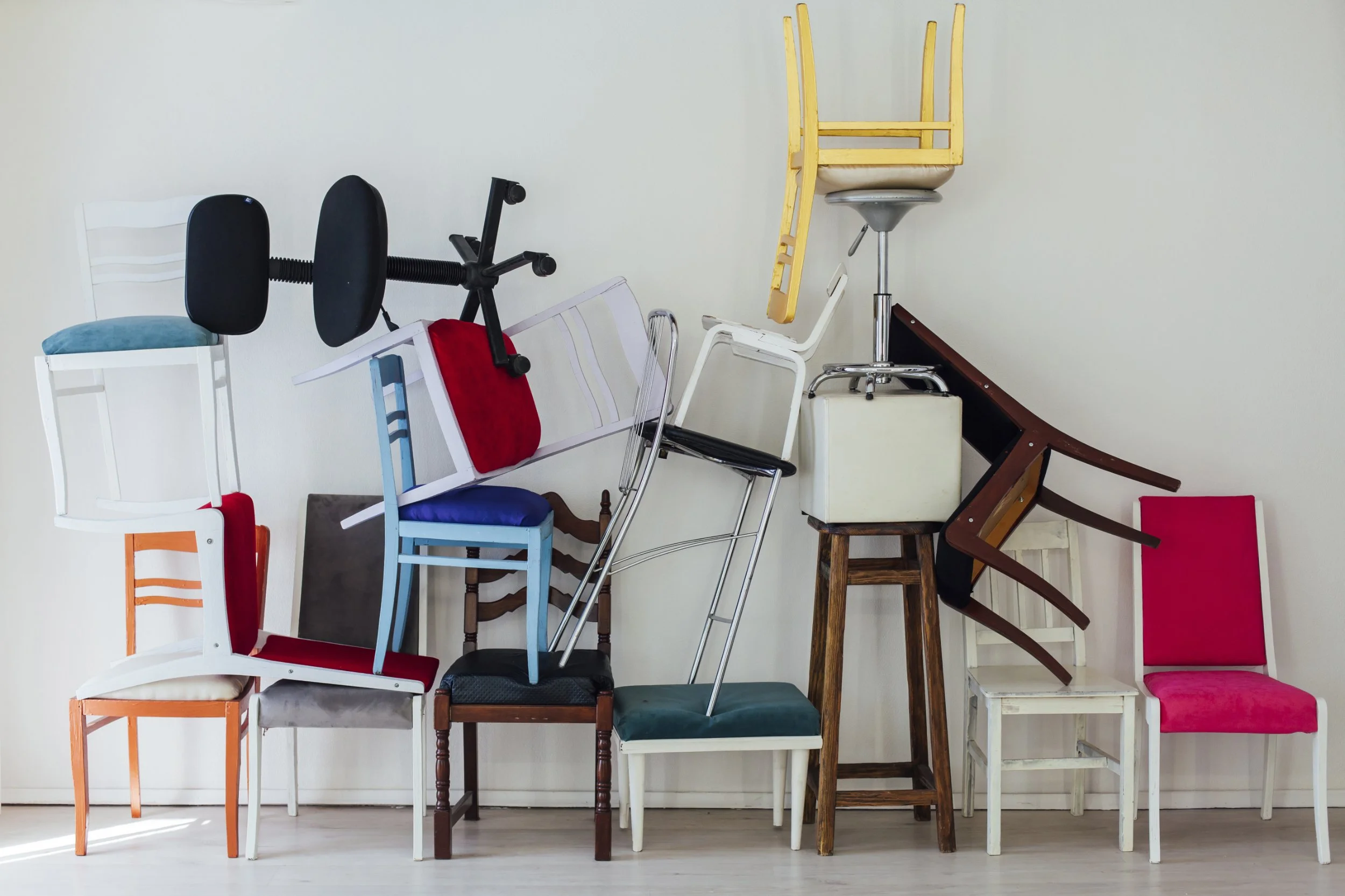 A pile of various chairs stacked and leaning against a plain white wall, including different styles, colors, and materials.