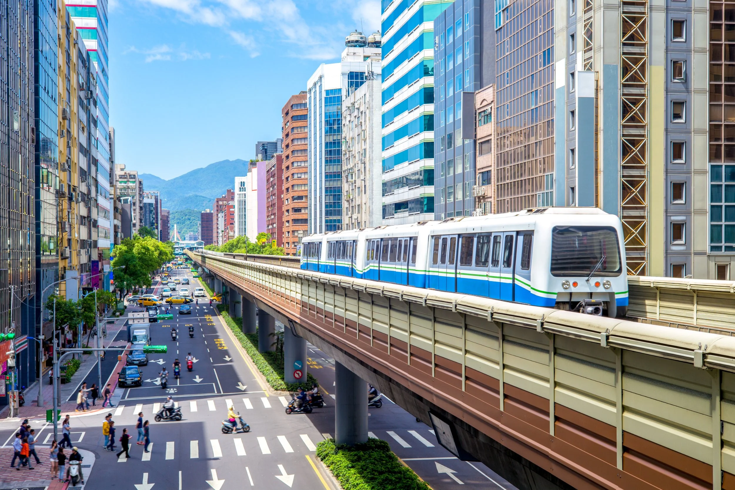 Urban scene with a train on an elevated track passing between tall modern buildings, busy street below with cars, motorcycles, and pedestrians, mountain range in the background.