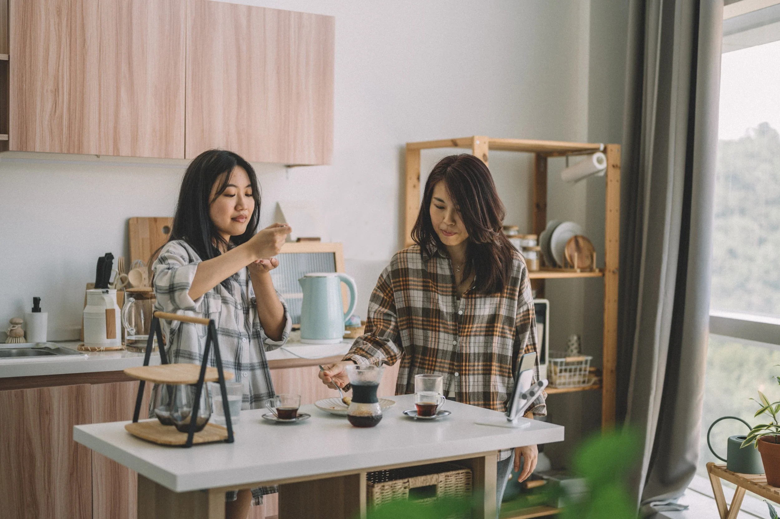 Two women in plaid shirts preparing coffee in a modern kitchen with wooden cabinets and a large window with curtains.