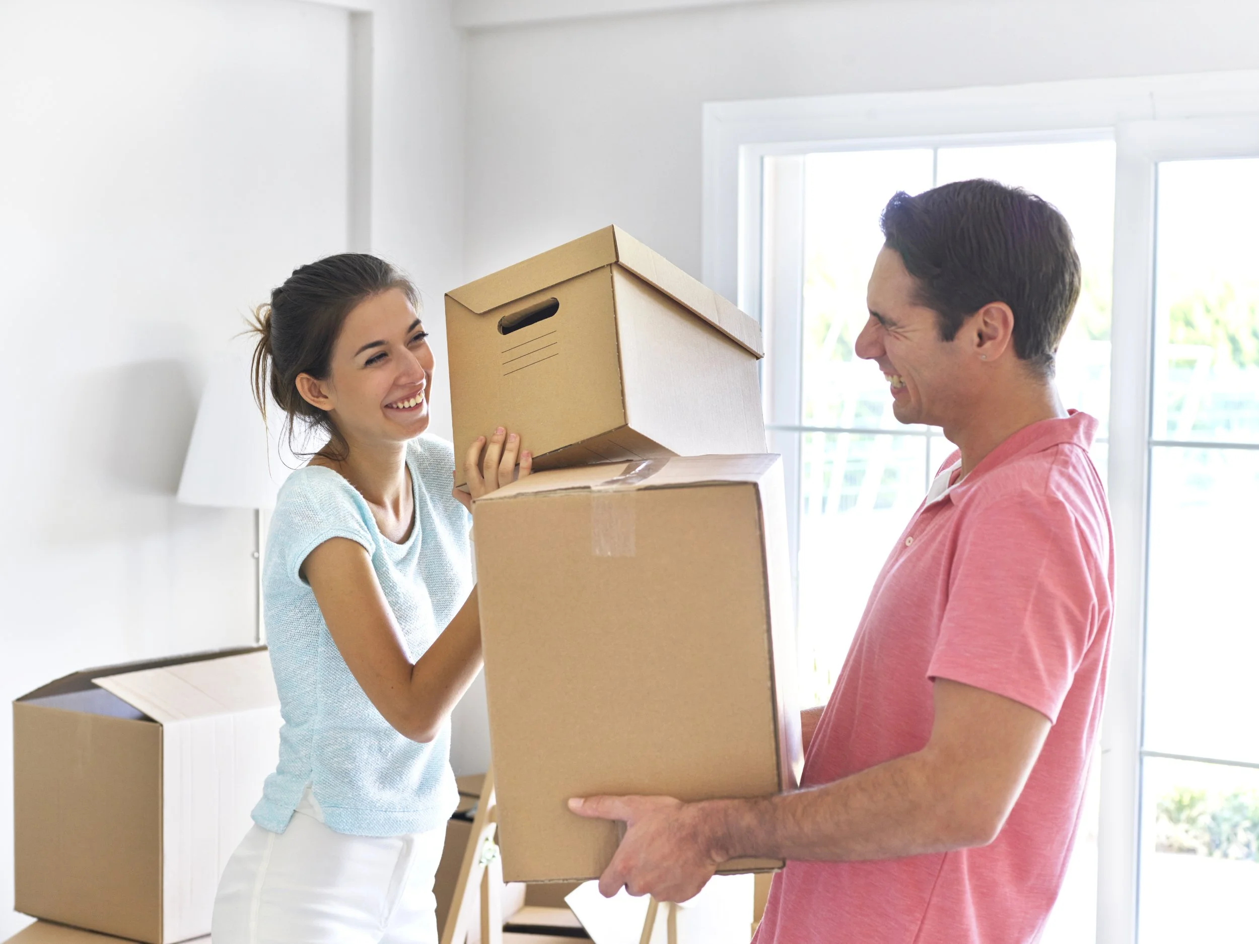 A young woman and a man smiling as they exchange boxes during a move in a bright room with large windows.