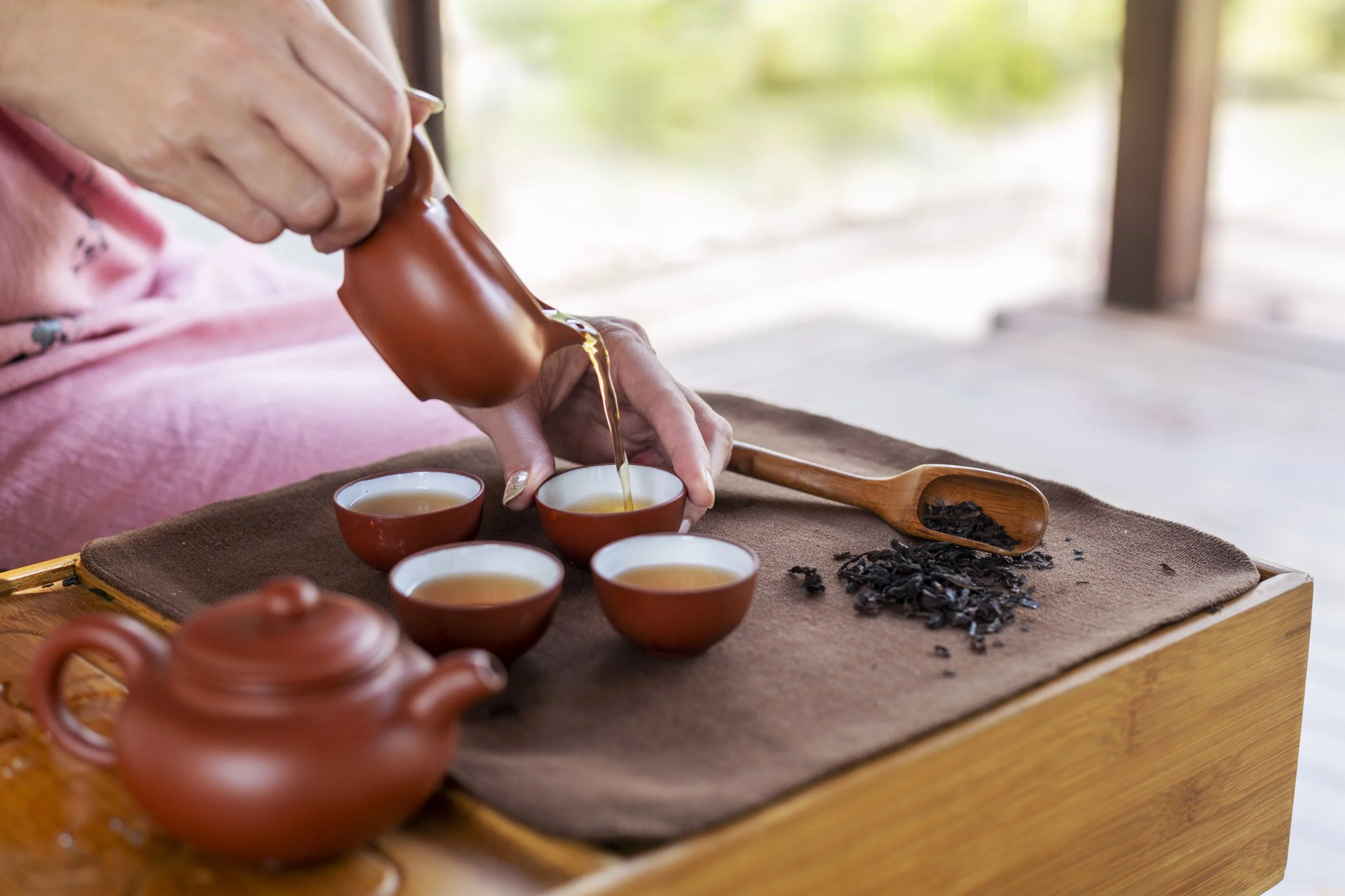 Person pouring hot tea from a brown teapot into small teacups on a wooden table, with loose tea leaves on a wooden scoop nearby.