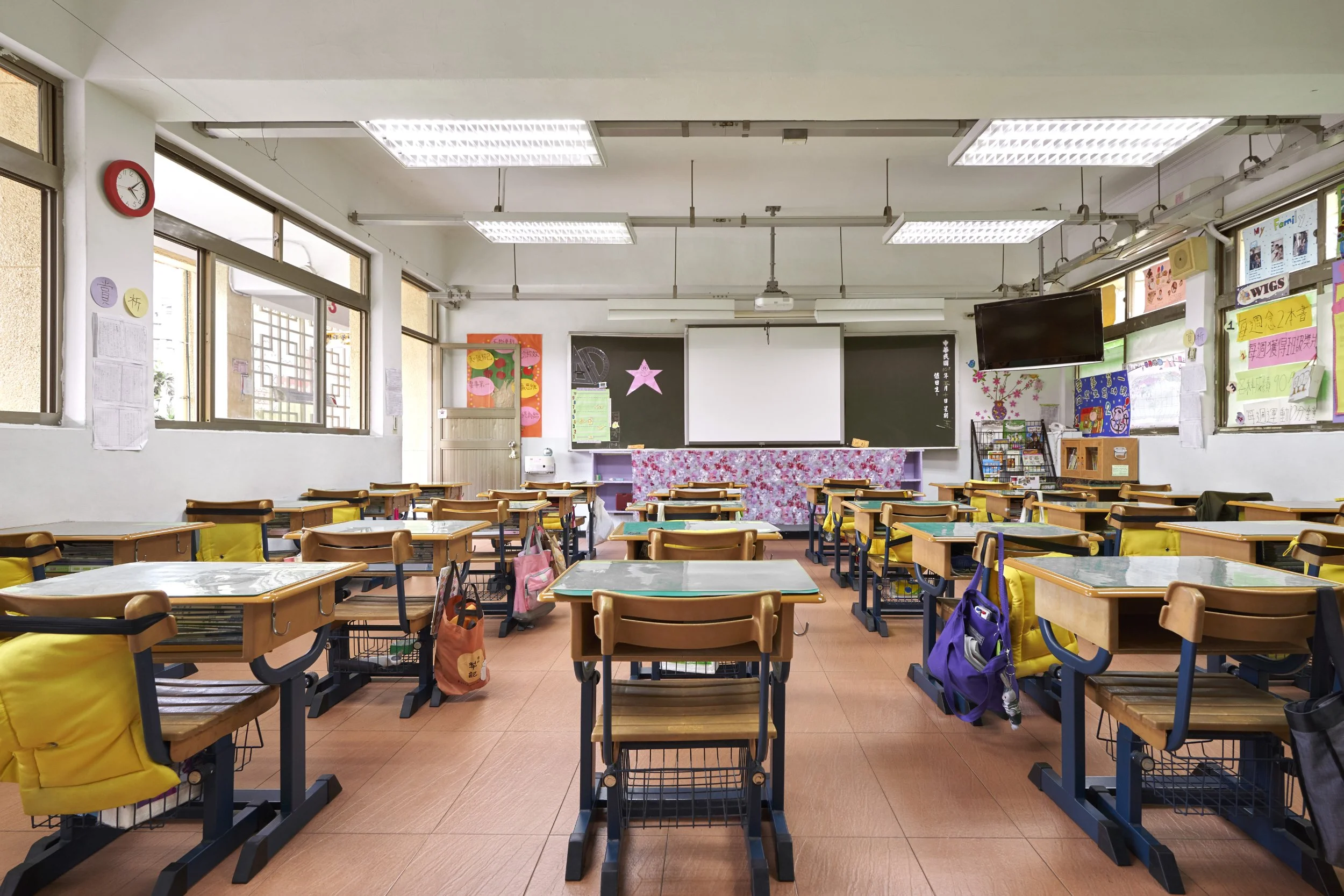 Empty classroom with desks and chairs arranged in rows, chalkboard at front with decorations, and colorful posters on walls.