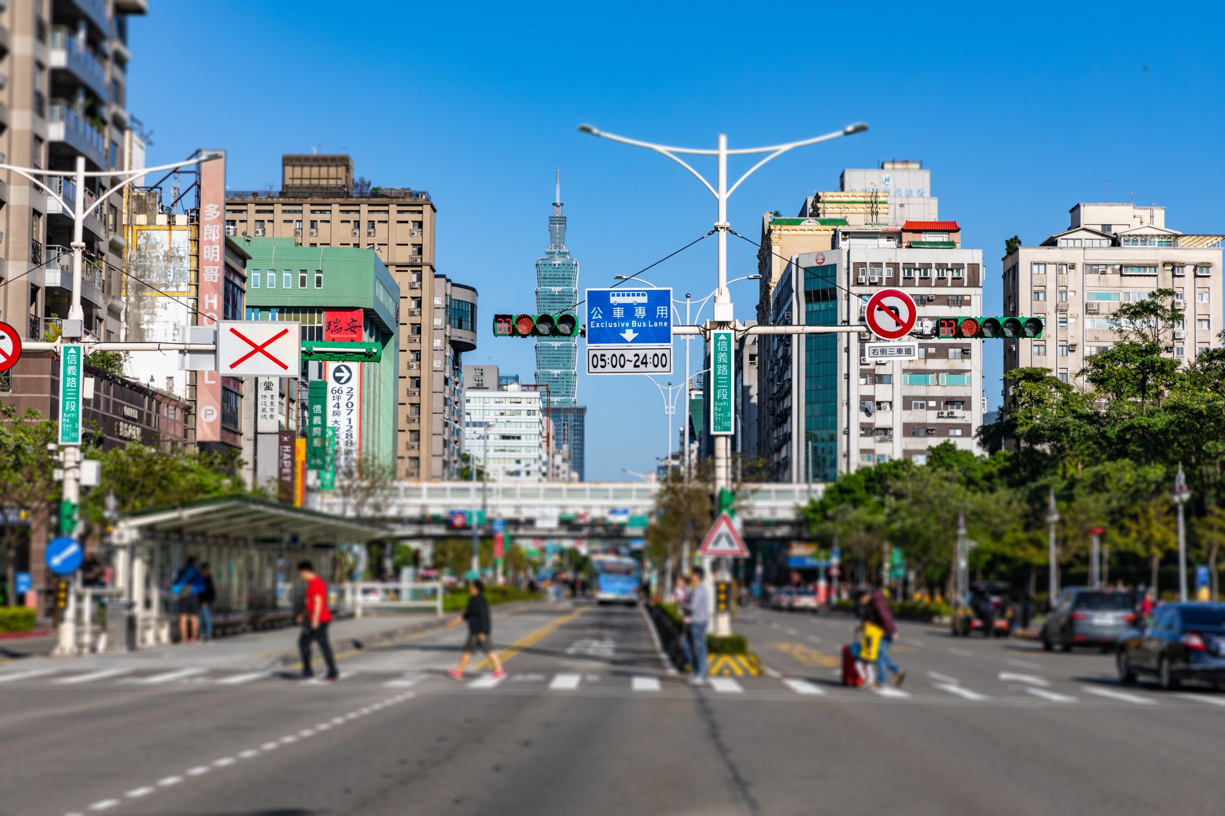 City street with pedestrians crossing at crosswalk, surrounded by tall buildings, road signs, traffic lights, and a clear blue sky. Taipei 101 Tower visible in background.