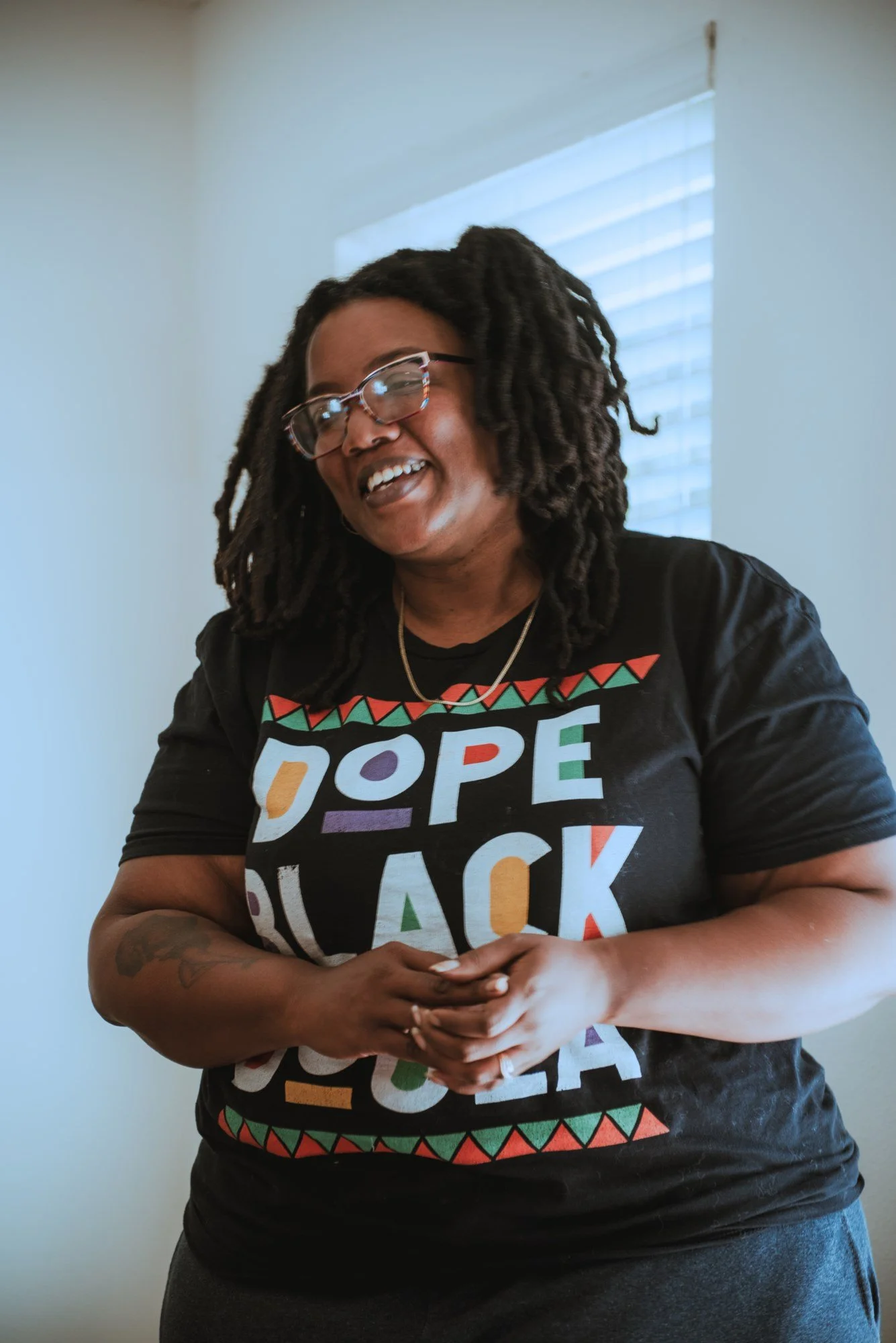 A woman with glasses smiling outdoors during sunset, with trees, houses, and pink flowering bushes in the background.