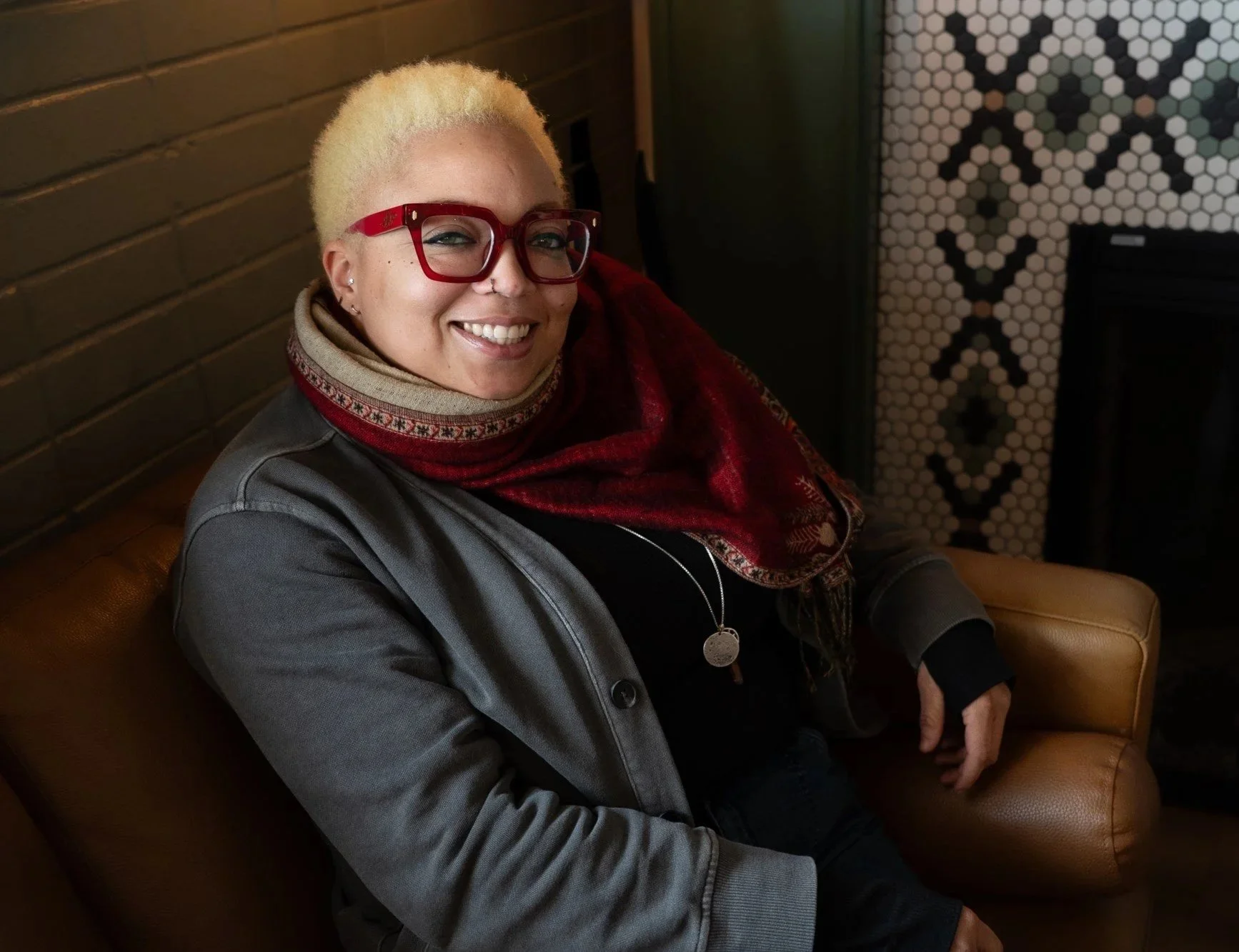 A smiling person with short dreadlocks, wearing clear glasses and a blue and white striped shirt, standing against a plain white wall.