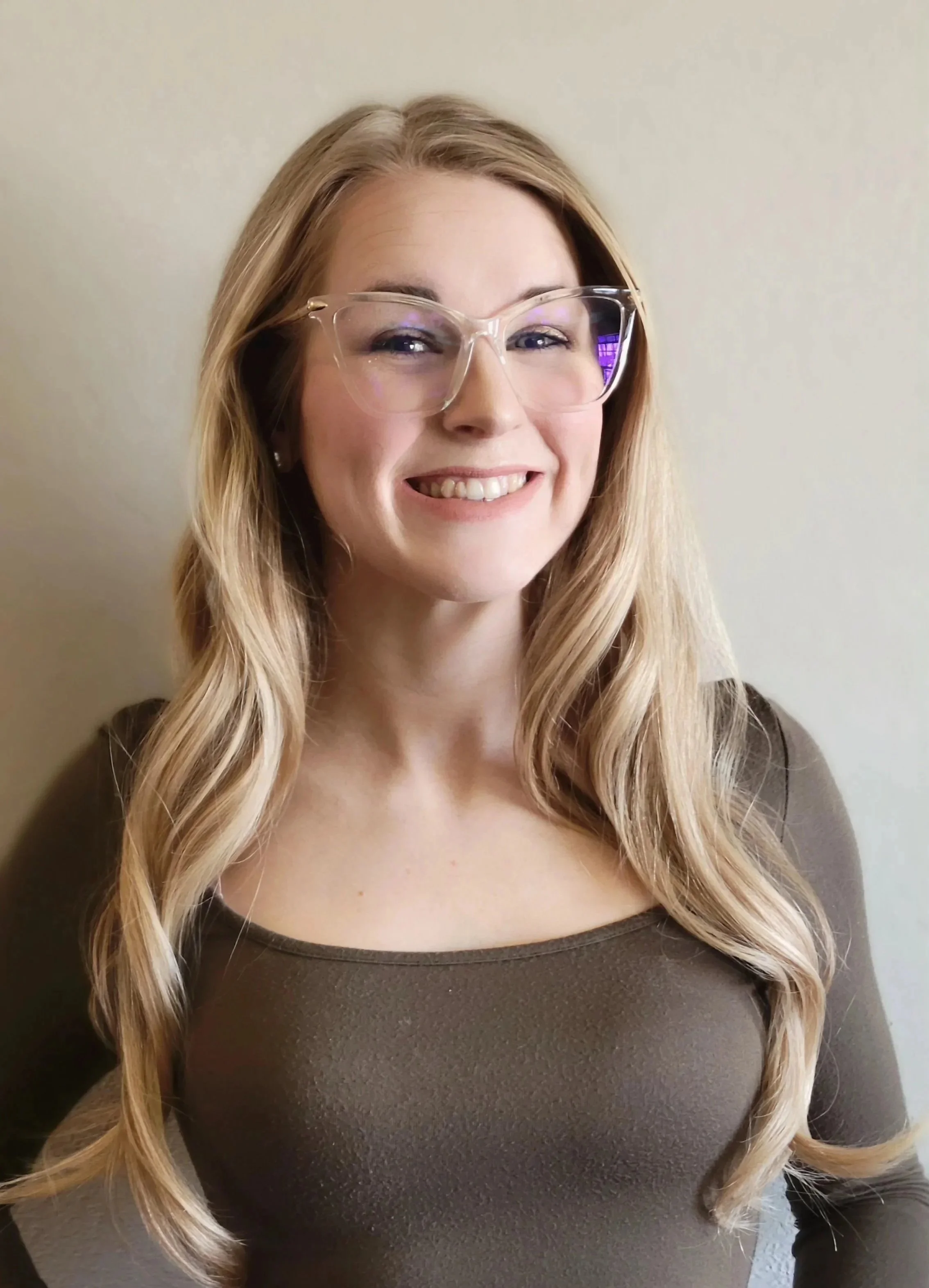A woman with long blonde hair wearing clear glasses and a brown top, smiling in front of a plain wall.