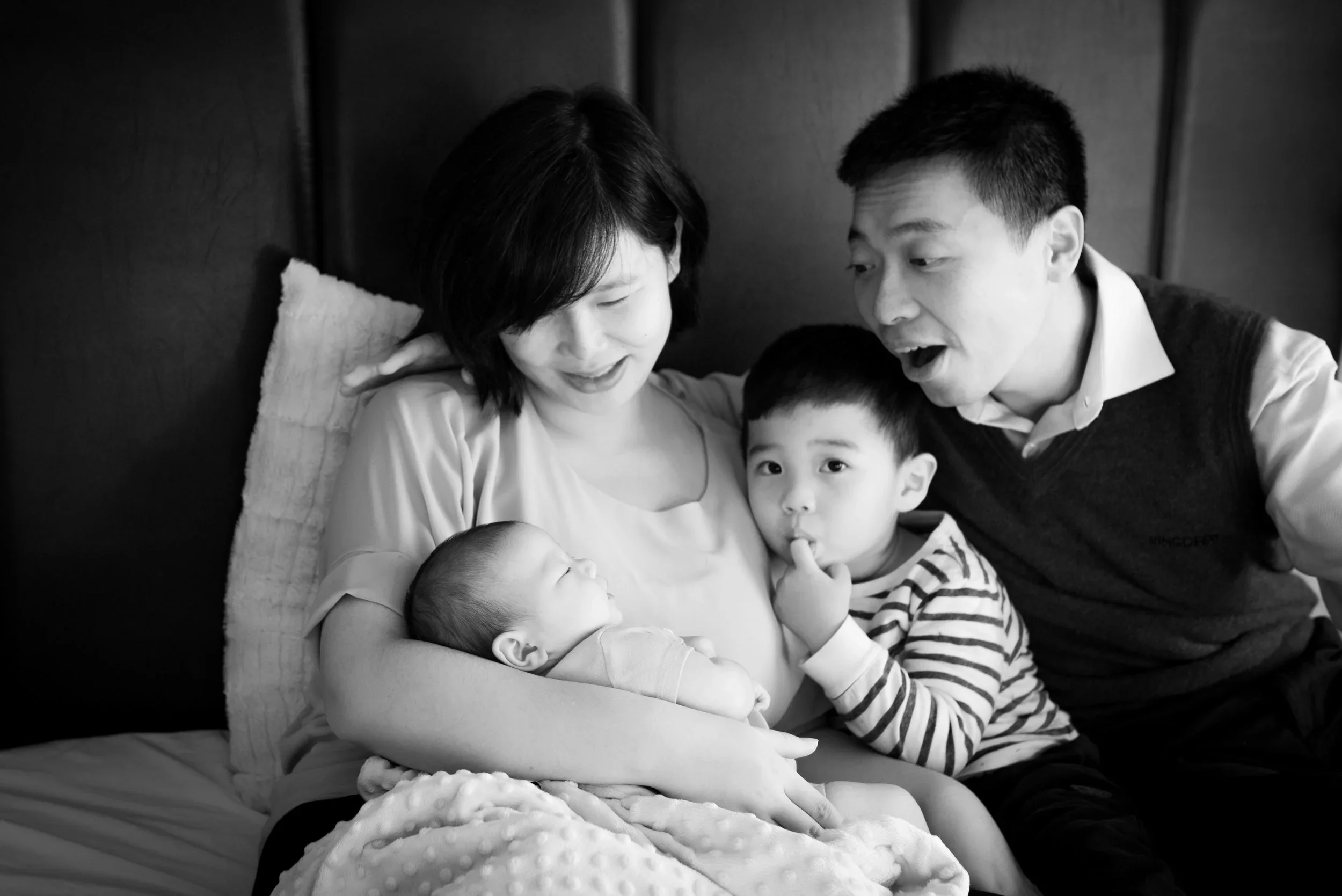 A family of four sitting on a bed, the mother holding a newborn, a young boy touching her face, and a father leaning in with an excited expression.