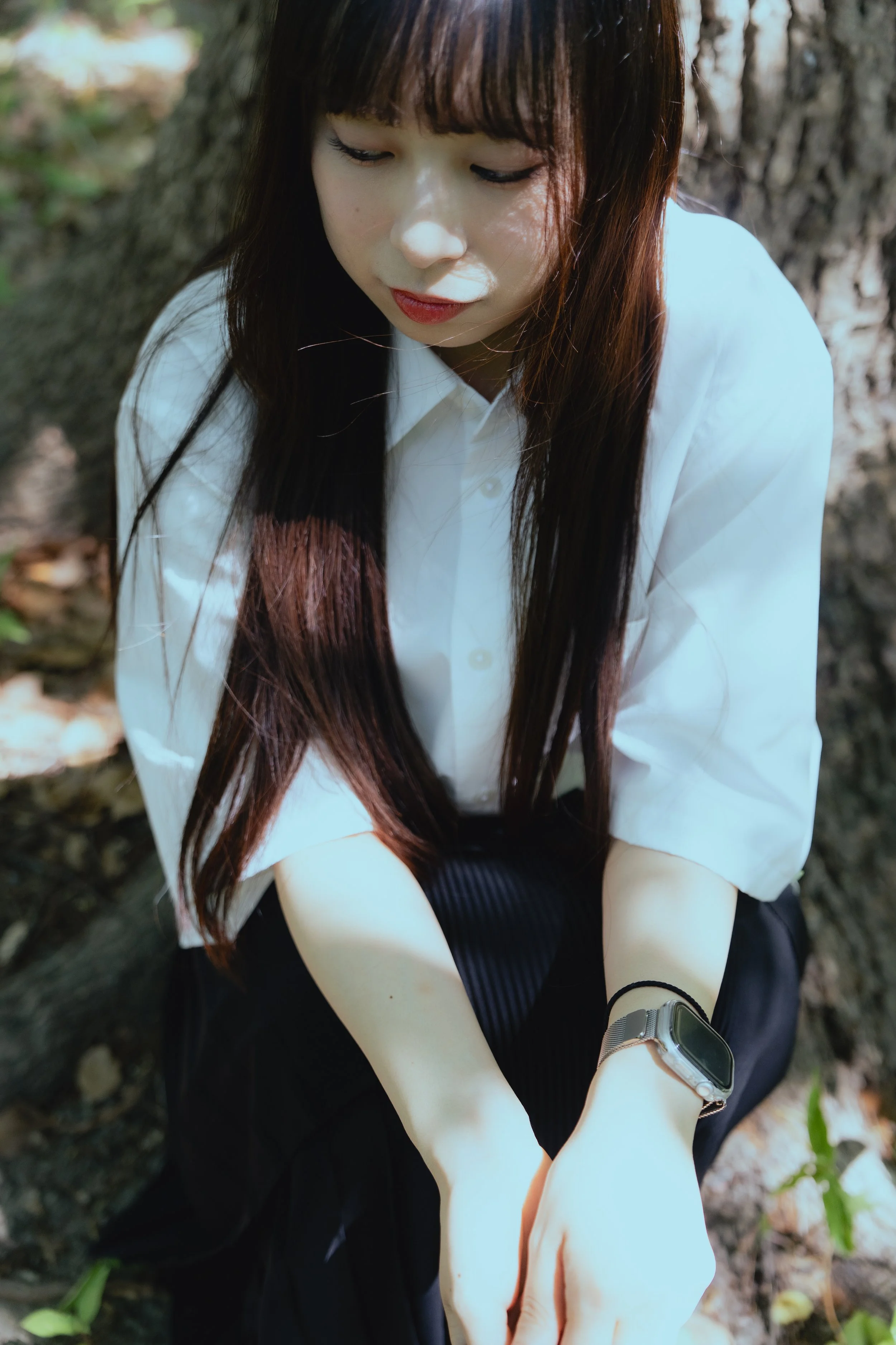 木の根元に座っている若い女性が、白いシャツと黒いスカートを着て、スマートウォッチを腕につけている。