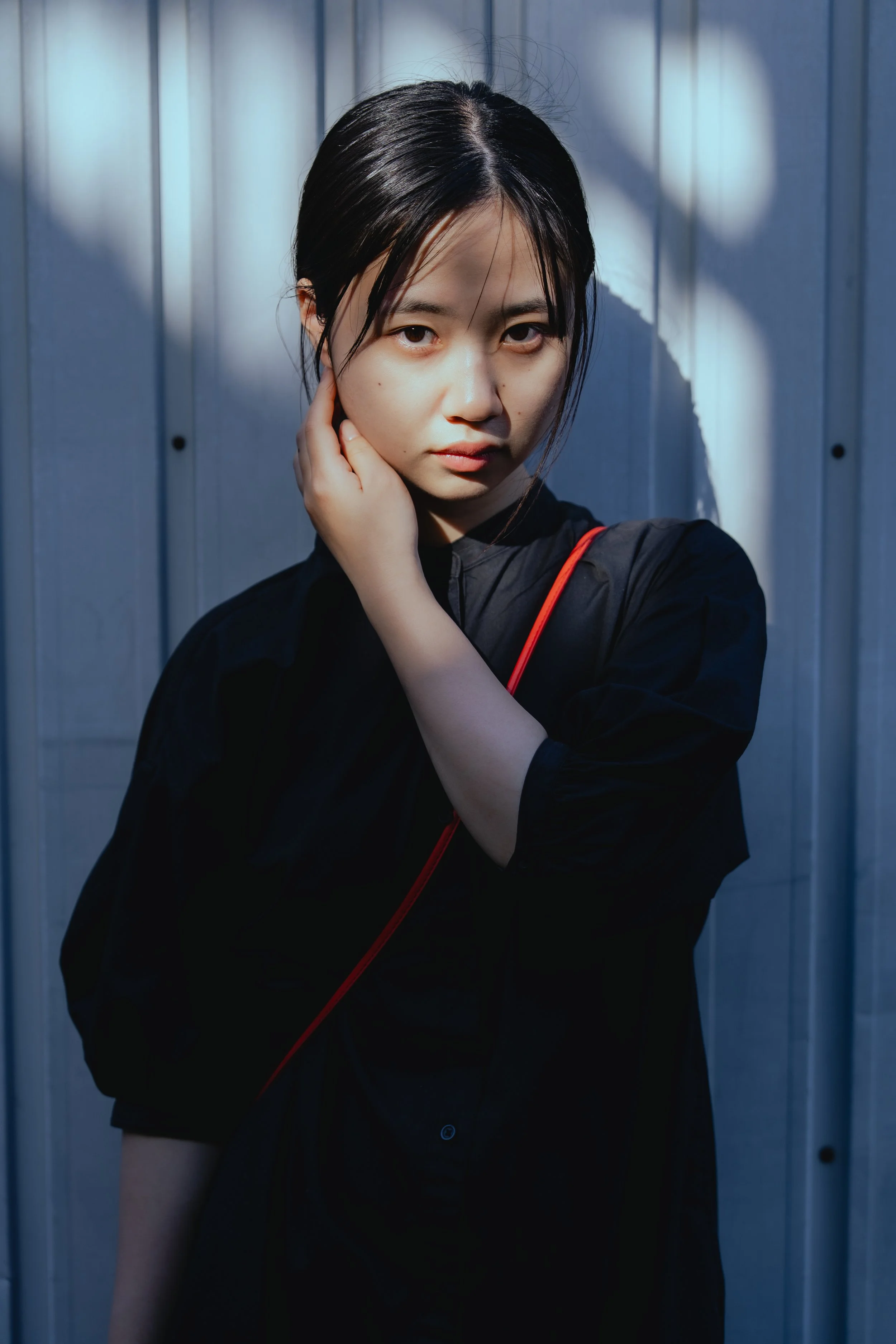 暗い背景で、黒いシャツを着た女性が真剣な表情でカメラを見つめているポートレート写真。彼女は右手を顔に近づけている。