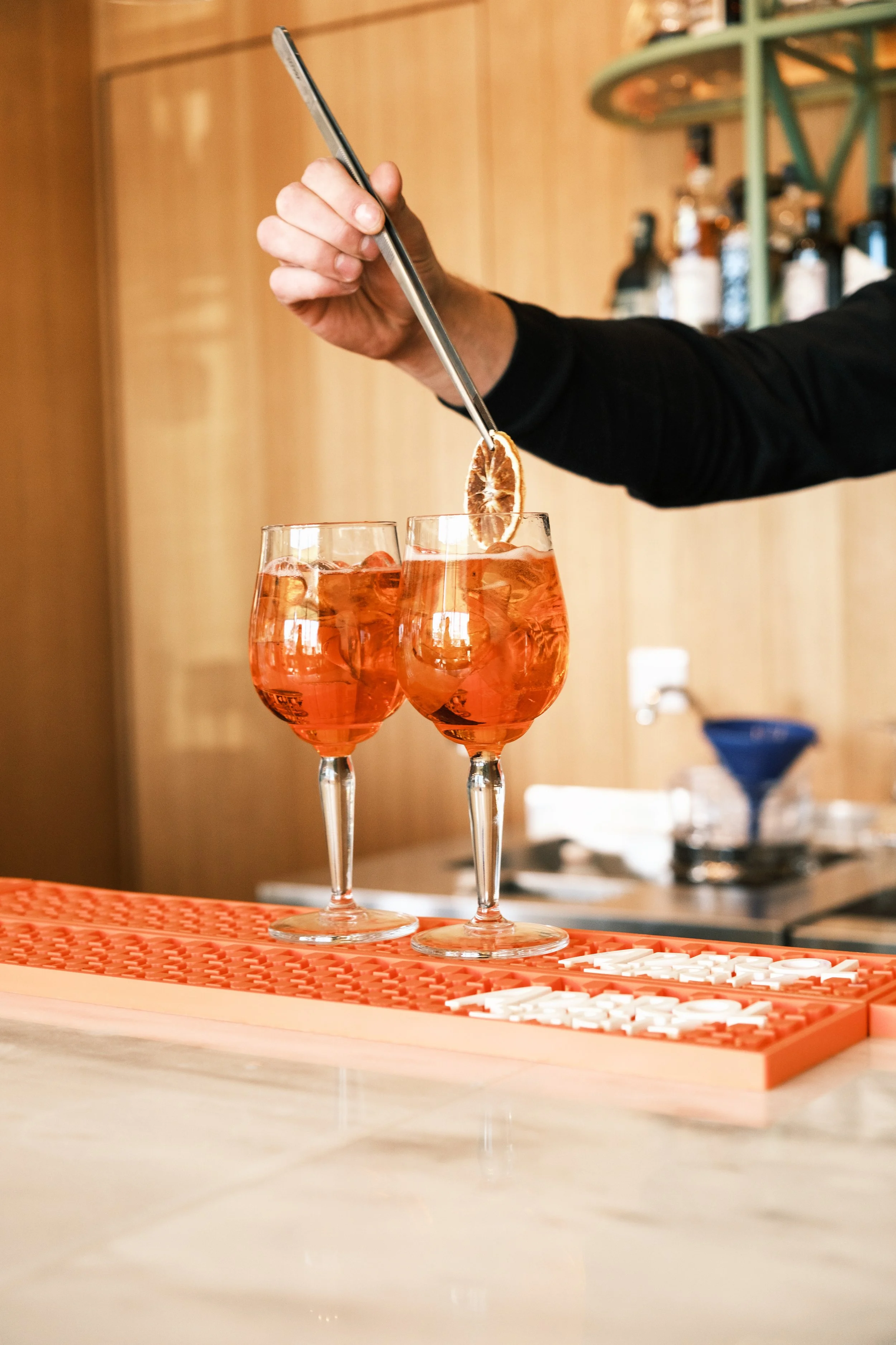 A bartender garnishing two glasses of pink aperitif with an orange slice using tongs.