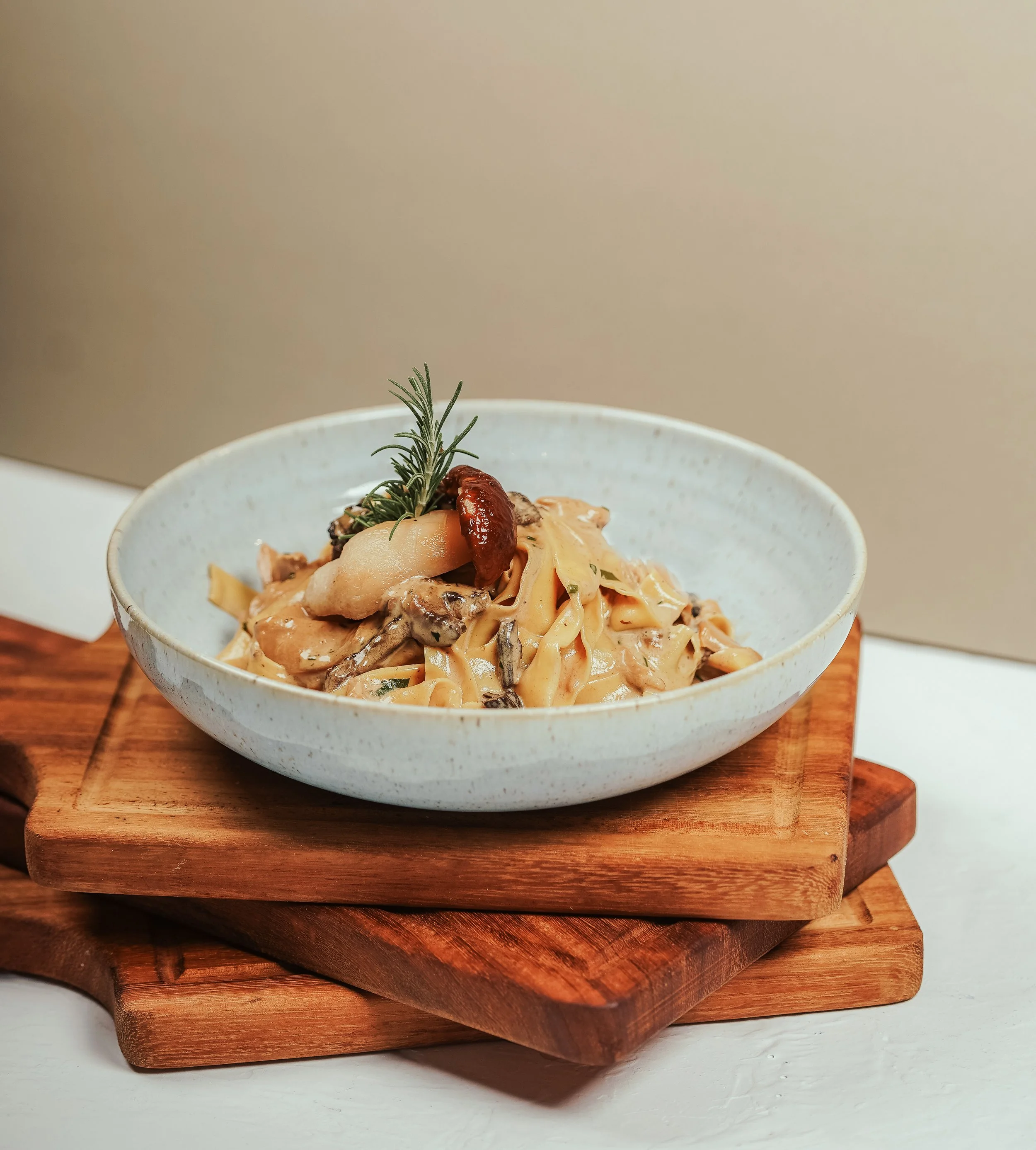 Creamy pasta with mushrooms, garnished with a sprig of rosemary and sun-dried tomato in a white bowl on wooden boards.