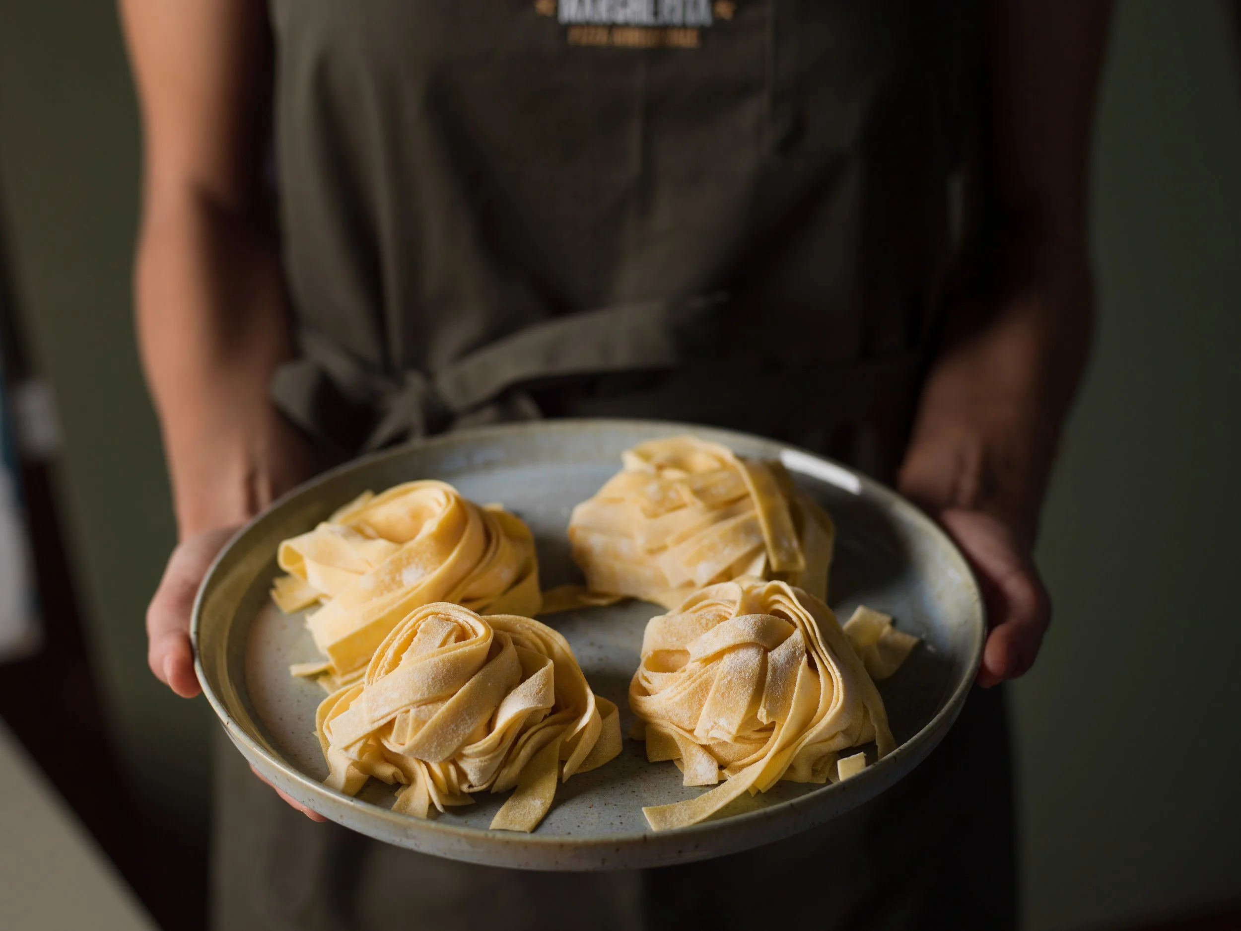 A person wearing a dark apron holding a round plate with five uncooked pasta nests.