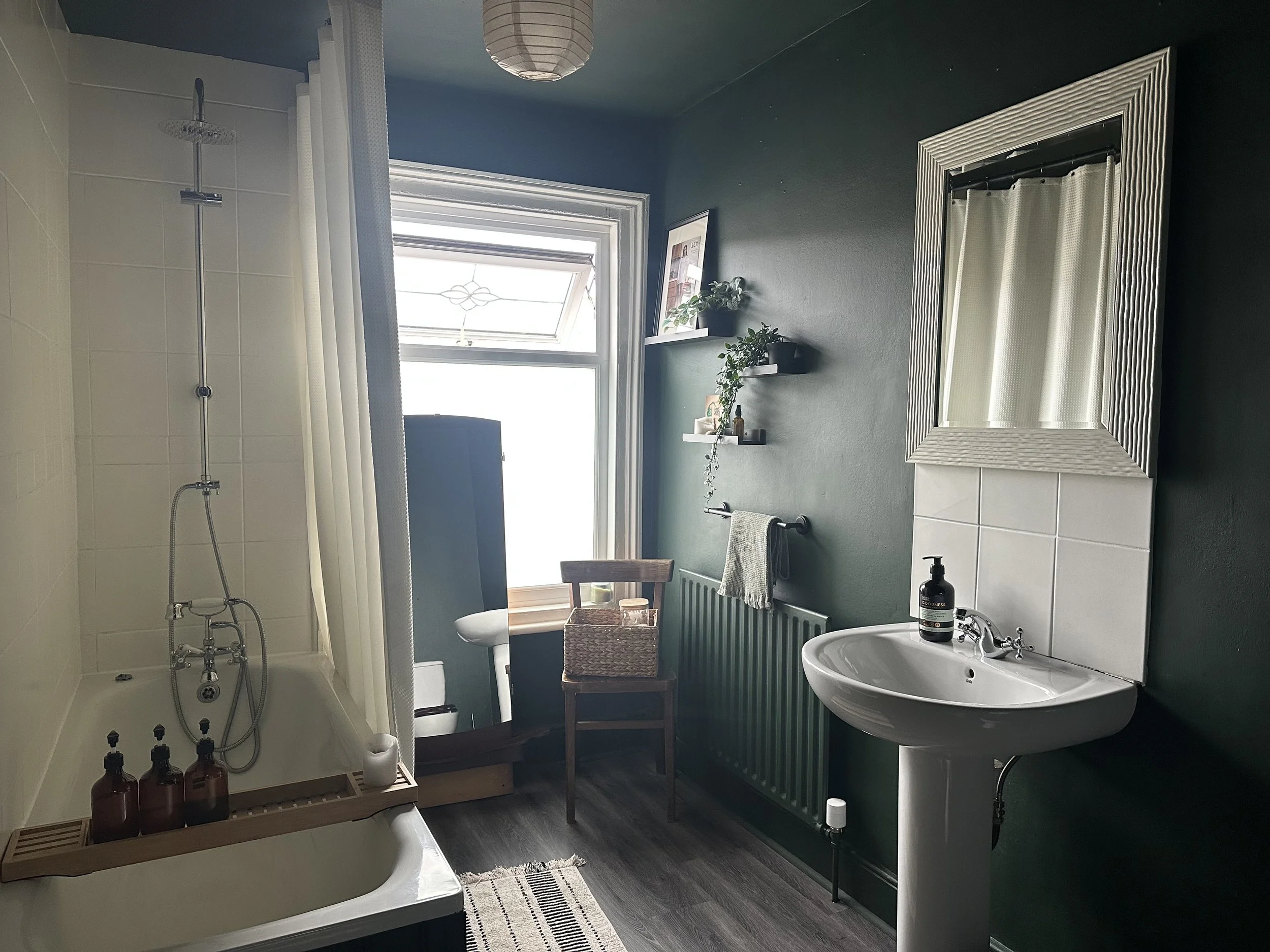 A bathroom with a shower area, a sink with a bottle of hand soap, a mirror, a radiator with a towel, a window with sunlight, a chair, and shelves with decorative items and plants.
