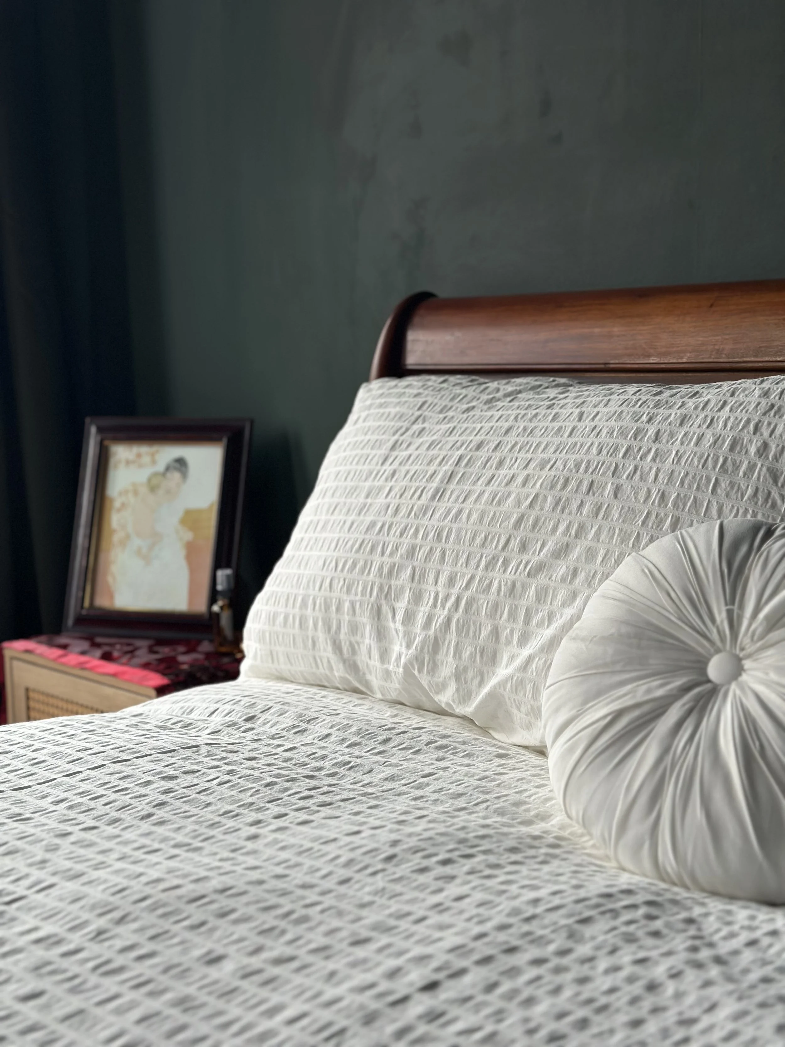 A neatly made bed with a white textured bedspread and a round, pleated pillow. In the background, a framed picture rests on a bedside table with a dark green wall behind.