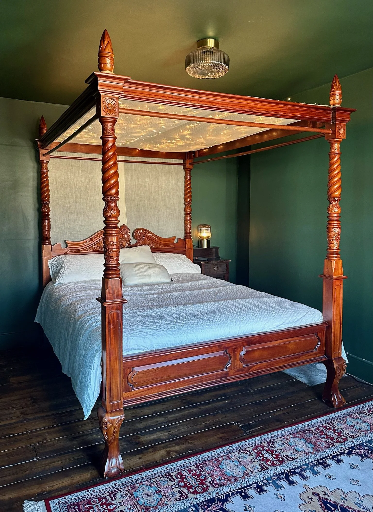 A wooden four-poster bed with carved posts and headboard, topped with a canopy decorated with fairy lights, in a room with green walls, dark hardwood floors, and a patterned rug.