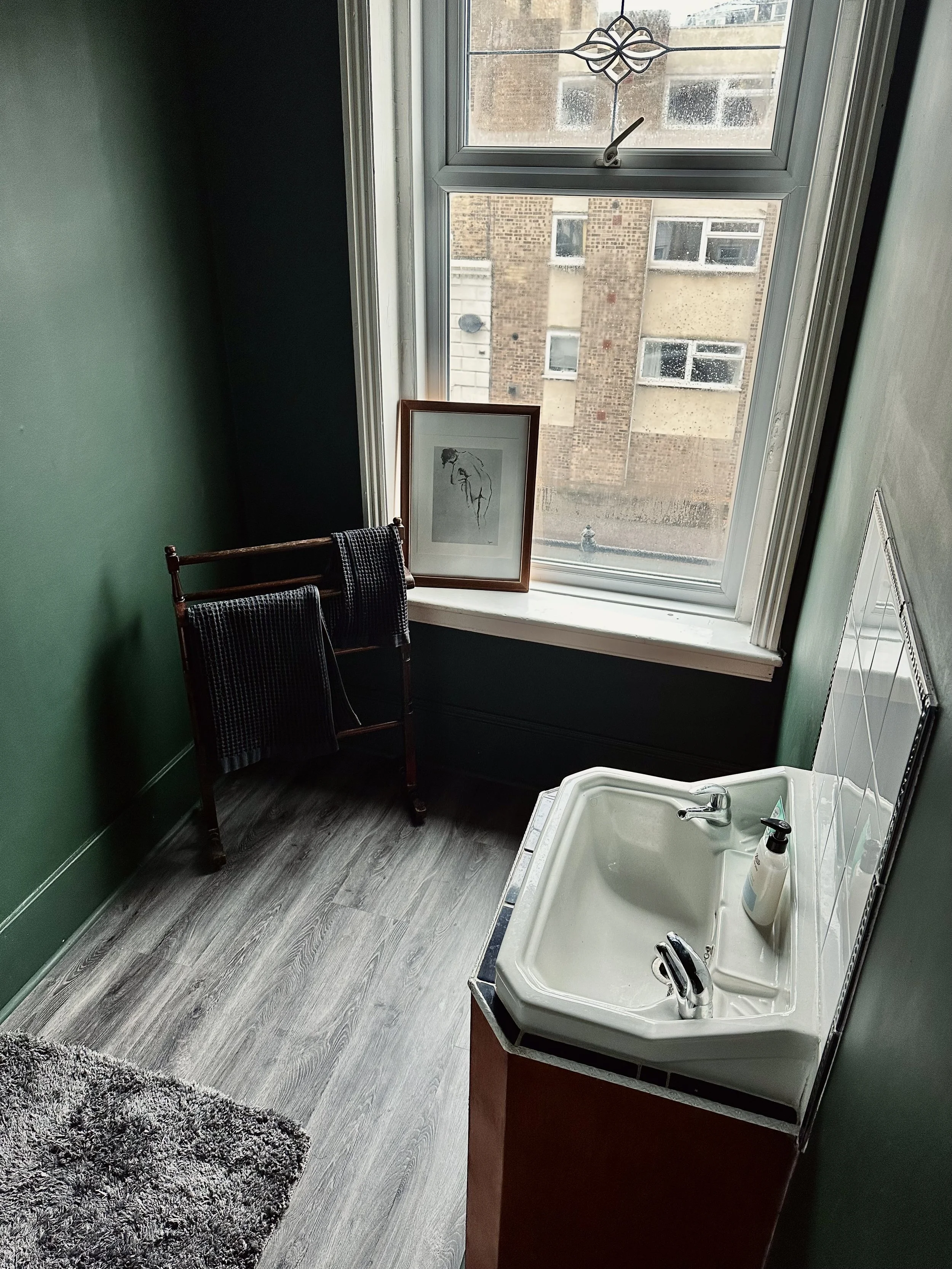 A small bathroom with green walls, a white sink with a soap dispenser, a framed sketch of a horse on the window sill, a textured gray rug, a black towel rack with two black towels, a large window with a view of an urban building, and a decorative wal