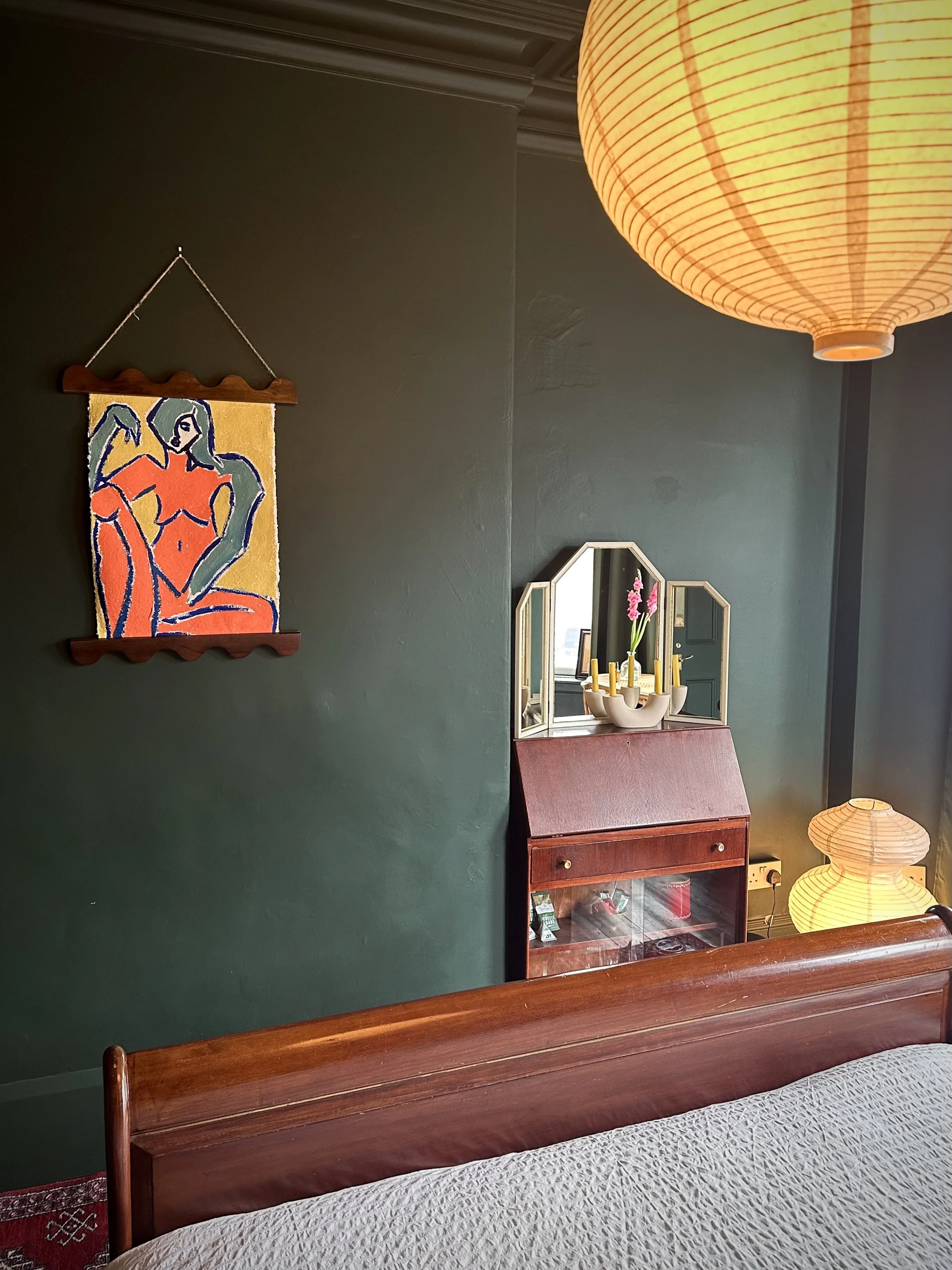 A bedroom wall painted dark green with a colorful abstract art piece hanging on it, a wooden dresser with a mirror, pink orchids in a white vase, and yellow candles. There is a large paper lantern ceiling light and a smaller one on the nightstand, wi