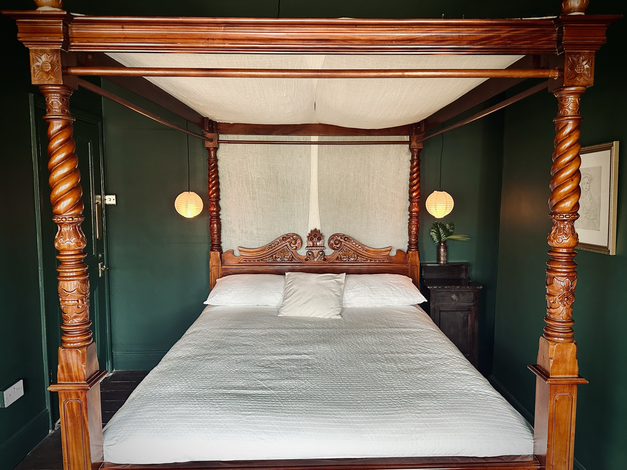 A four-poster wooden bed with white bedding and pillows, flanked by a dark bedside table with a potted plant, in a room with dark green walls and hanging paper lantern lights.