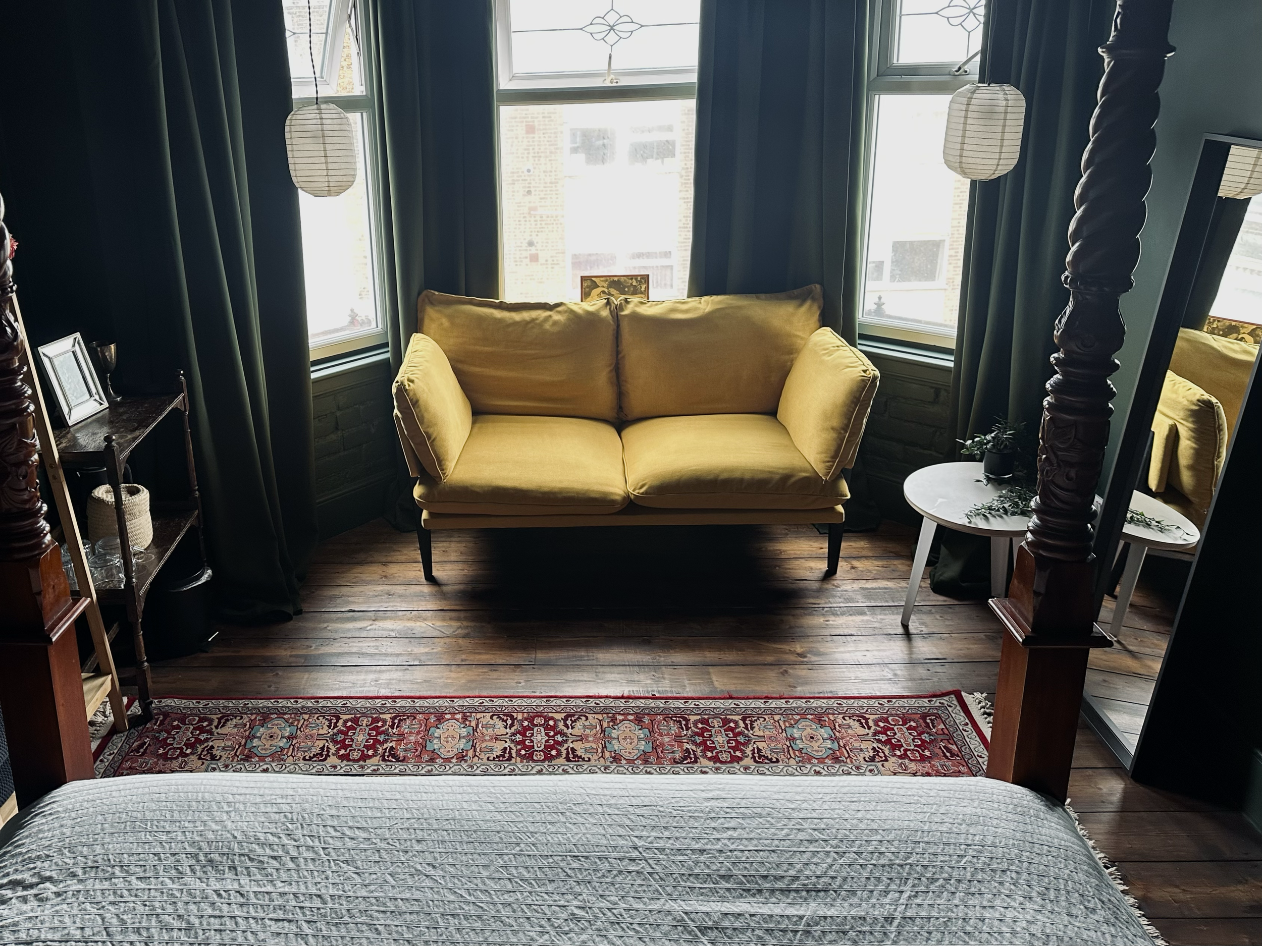 A yellow sofa in a room with dark green walls, three large windows, dark curtains, a small white side table with a plant, a tall mirror, and wooden flooring with a patterned rug.