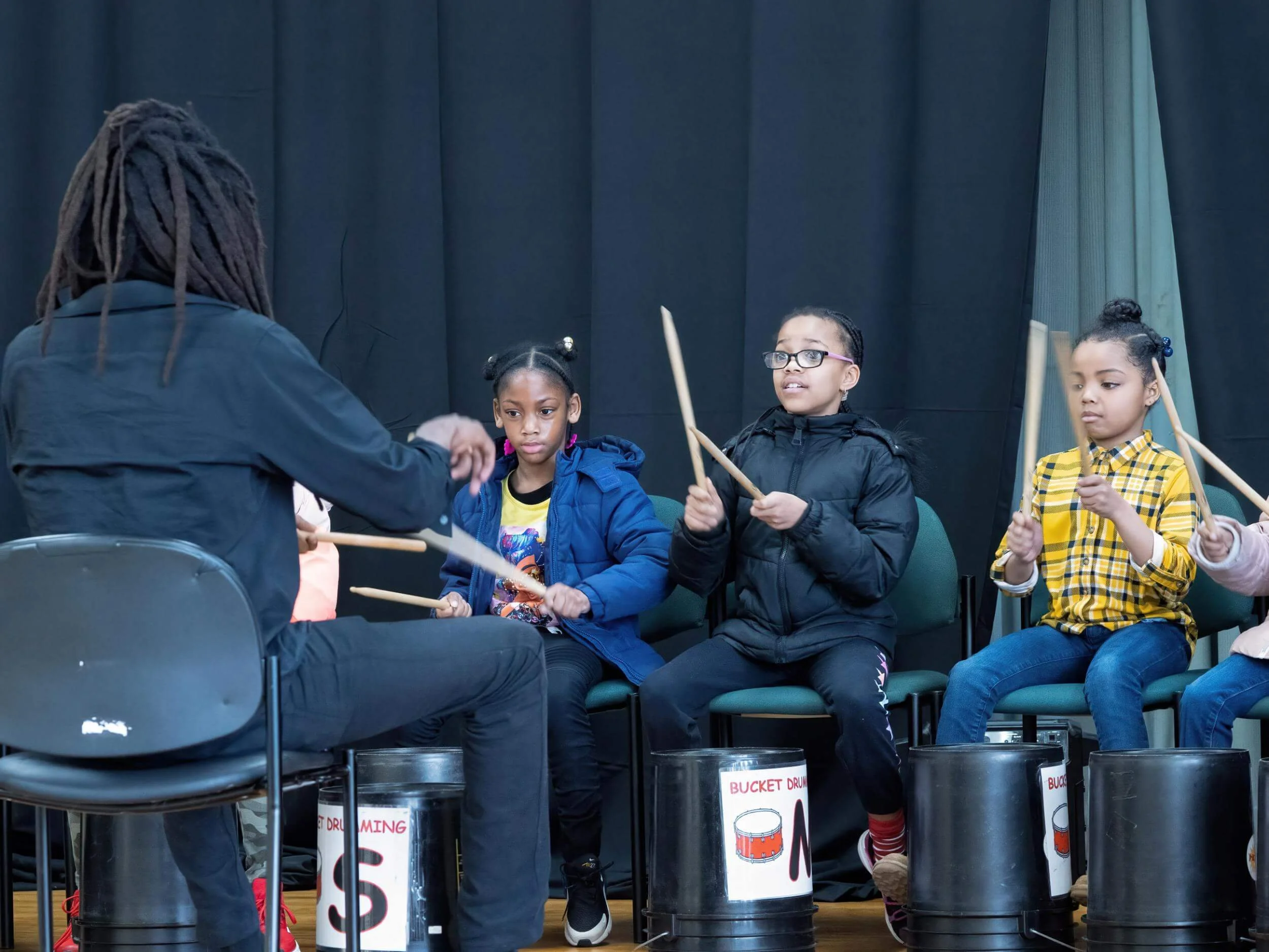 Children sitting on chairs playing drums led by an instructor during a music class.