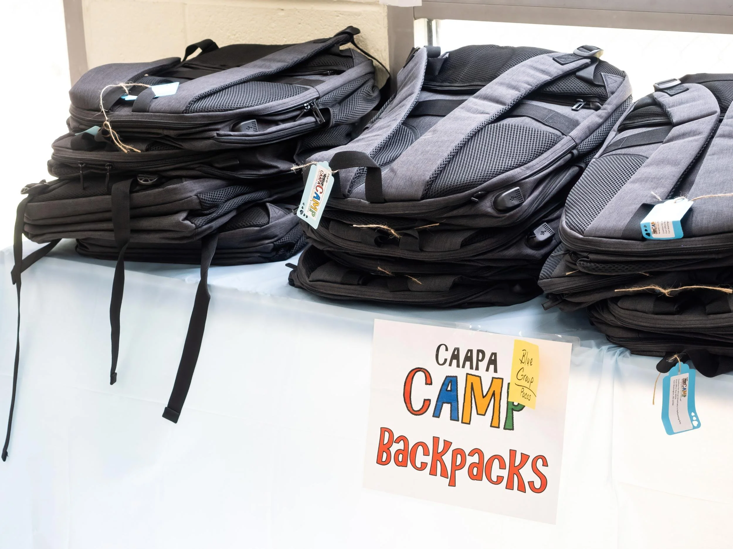 Stacks of black and gray backpacks on a table with a sign that reads 'Camp Backpacks'.