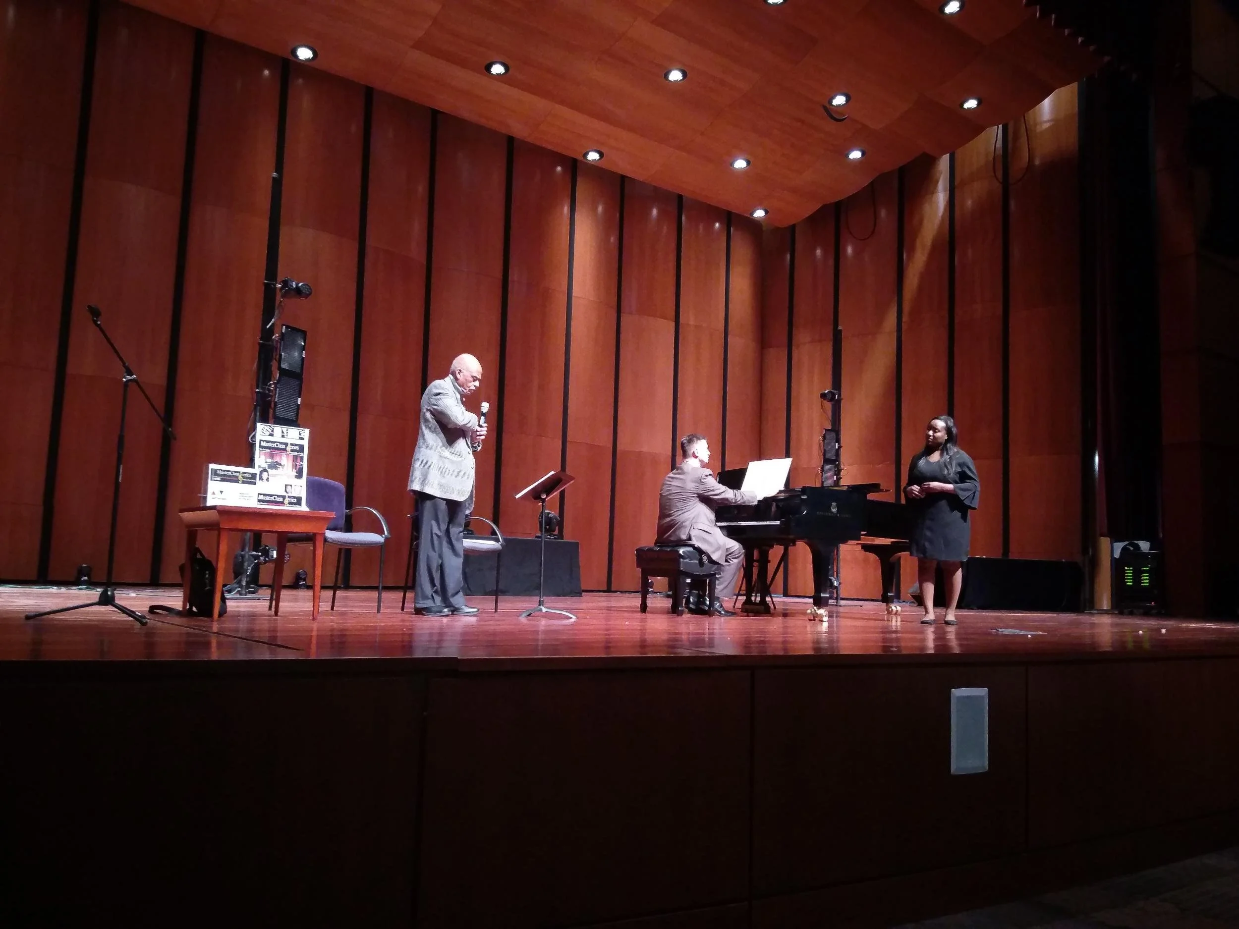 A musical performance on stage with a man singing, a woman singing, and a man playing the piano in a wood-paneled concert hall.