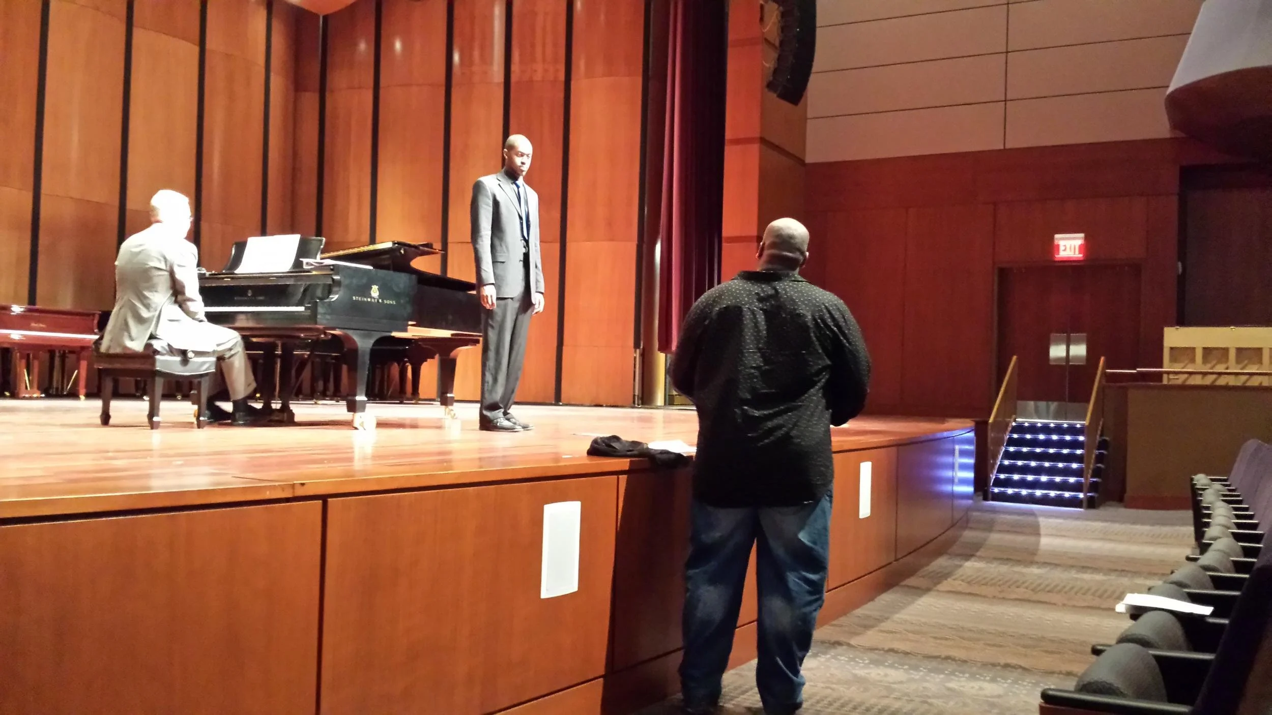 A person in a black shirt and jeans standing in front of a stage with a pianist and a singer rehearsing.