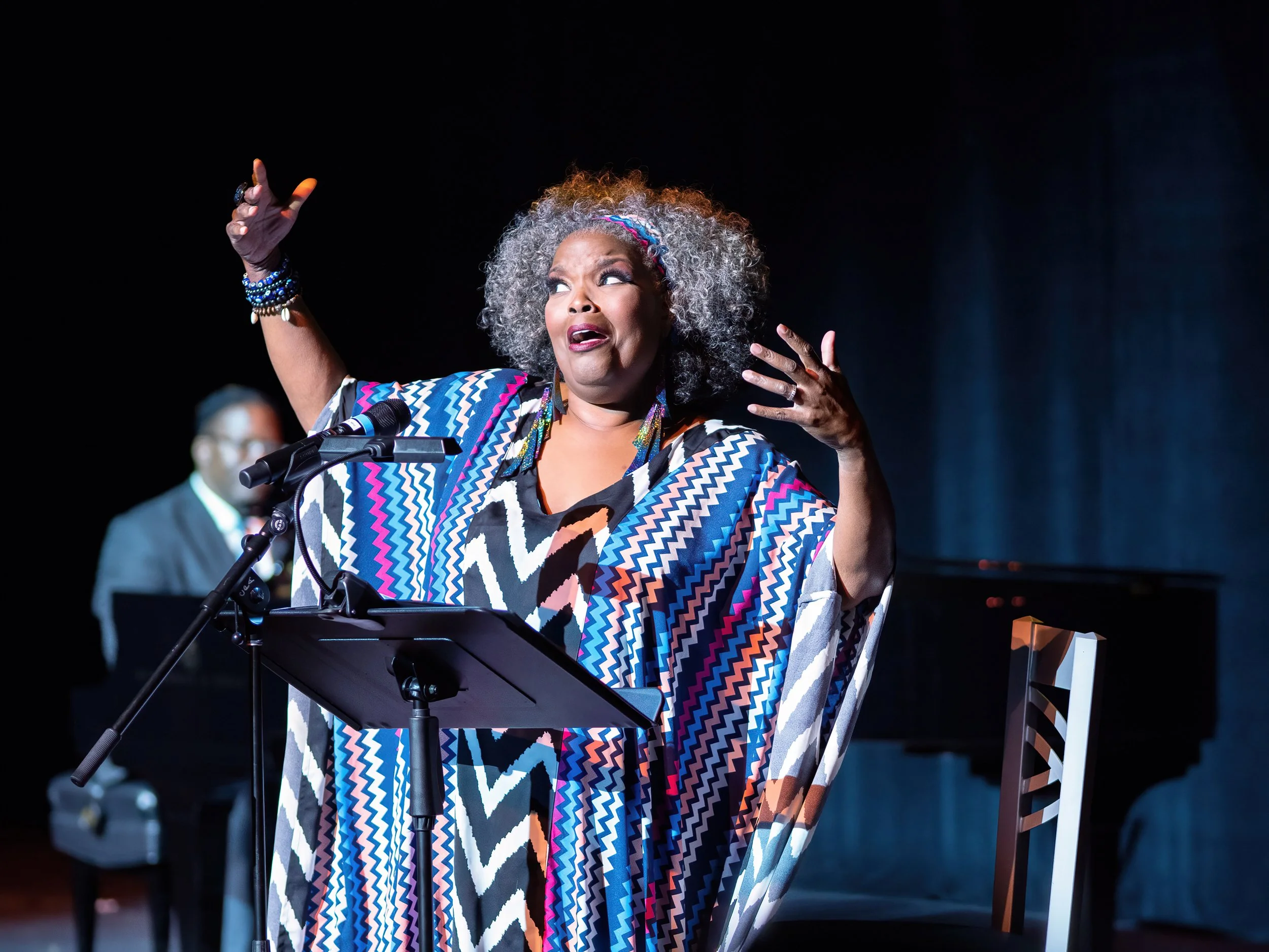 A woman with curly gray hair singing or speaking passionately at a microphone on stage, wearing a colorful zigzag patterned dress, with a man playing the piano in the background.