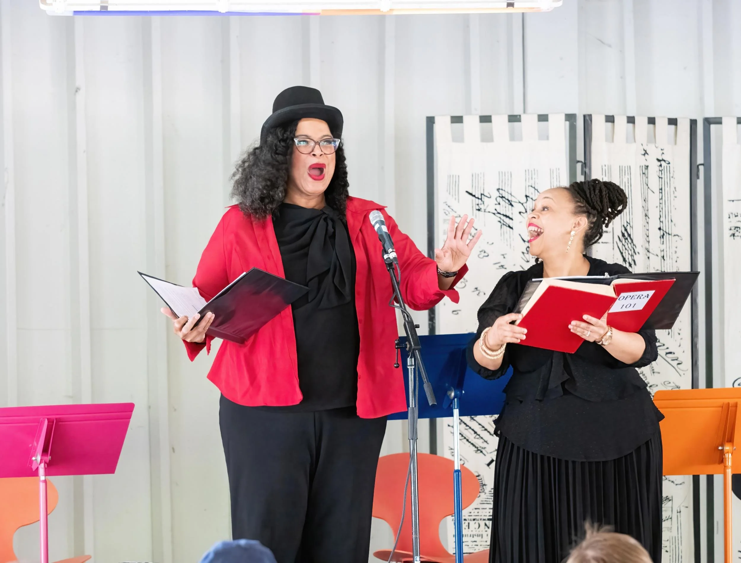 Two women holding open books, one talking and the other smiling, on a stage with music stands and sheet music in the background.