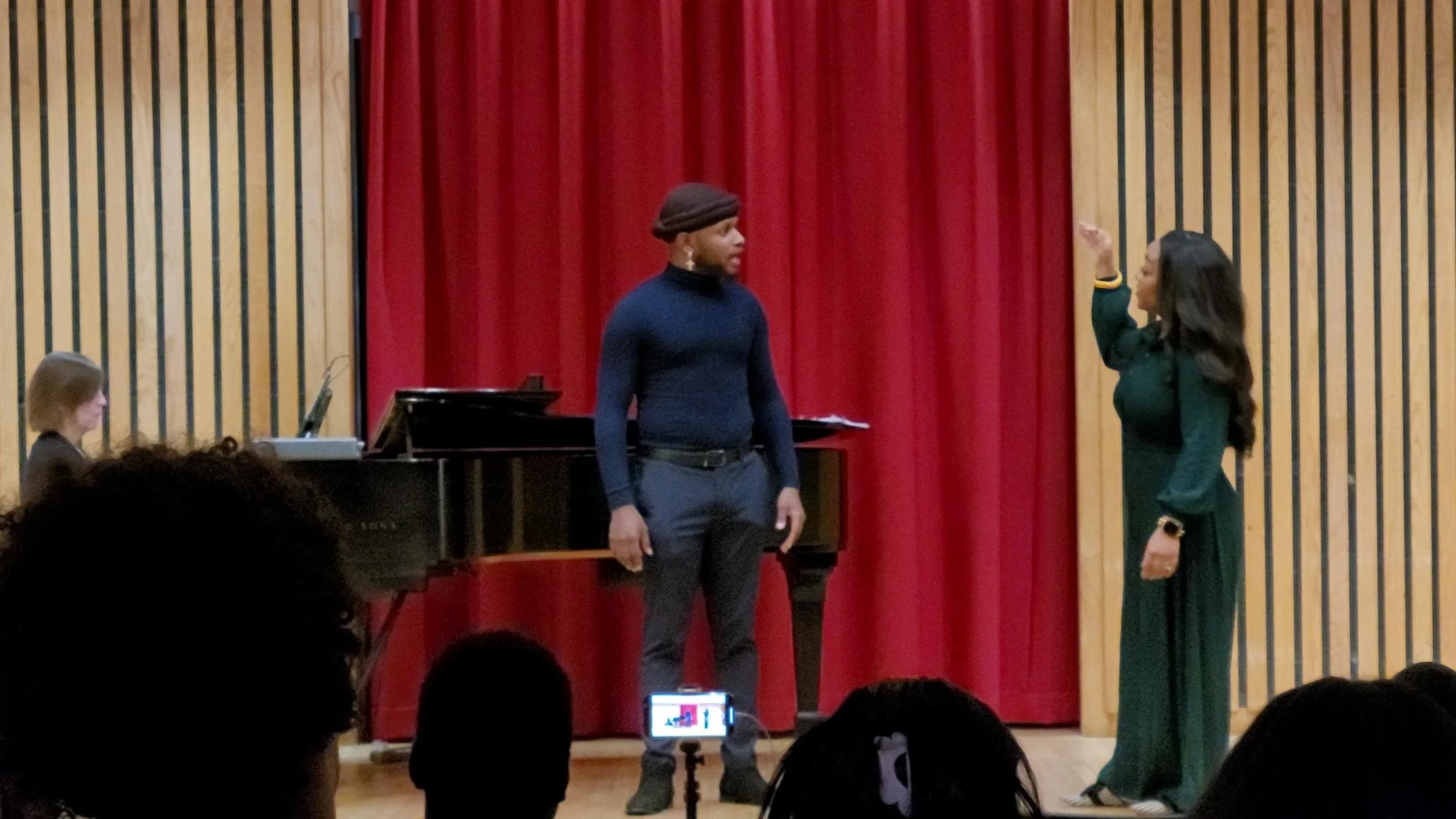 A woman in a green dress and a smartwatch appears to be conducting or instructing a man in a blue turtleneck and dress pants, with a black piano and a pianist visible in the background, on a stage with red curtains and wooden paneling.