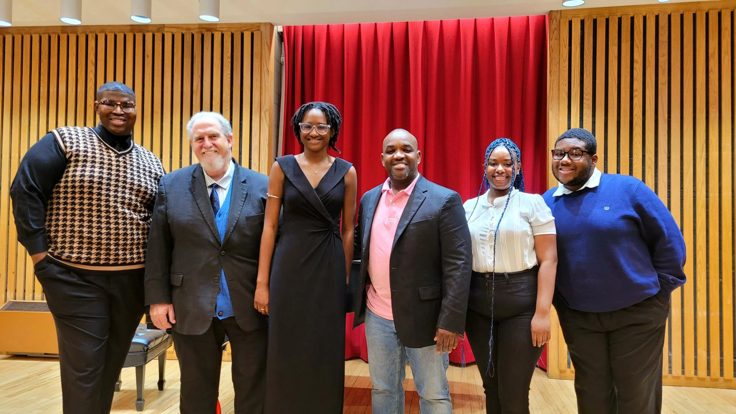 Group of seven diverse people standing together on a stage in front of red curtains, smiling for a photo.