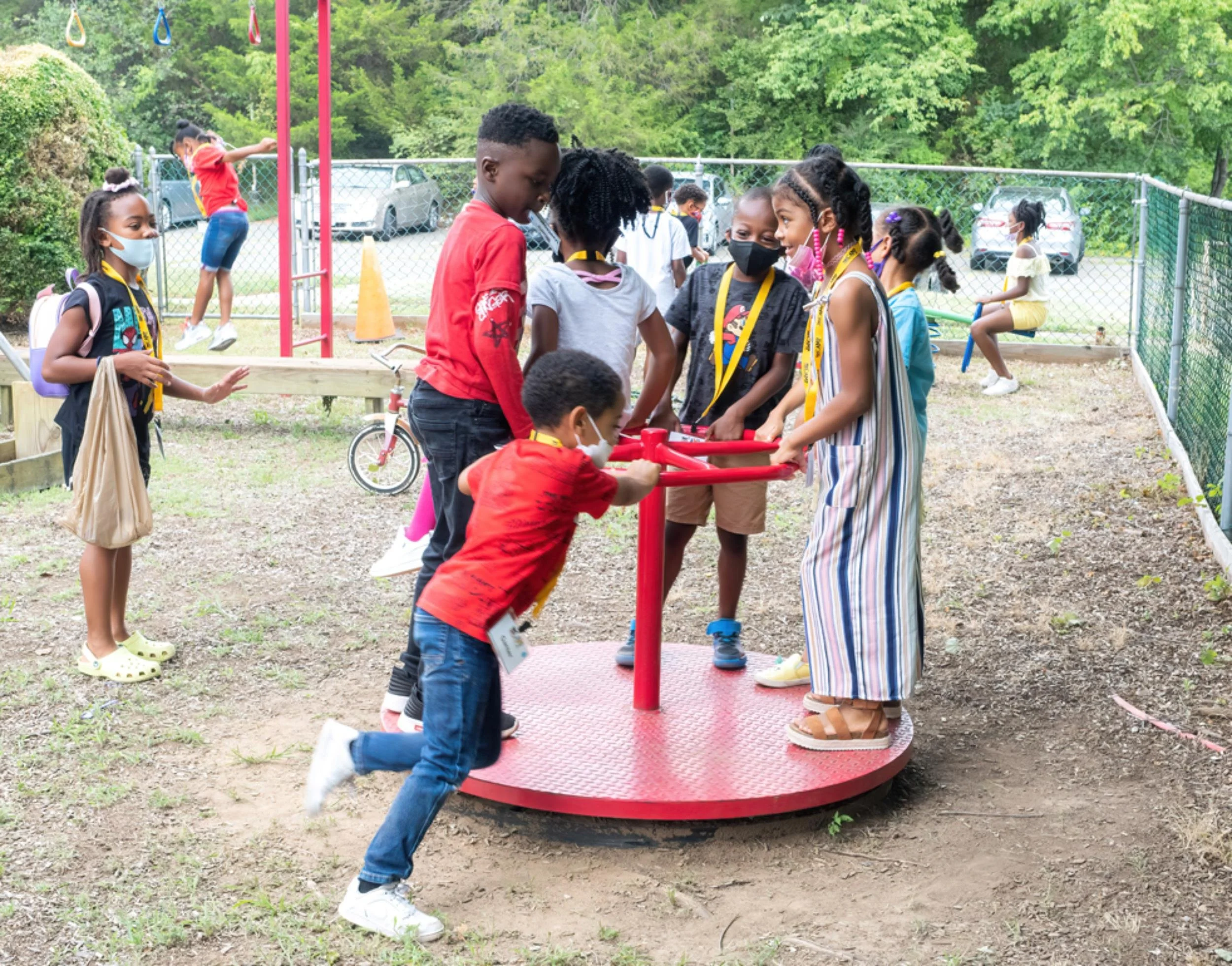 Children playing on a red spinning playground equipment outdoors, with several kids waiting and some playing in the background, wearing masks and casual clothing.