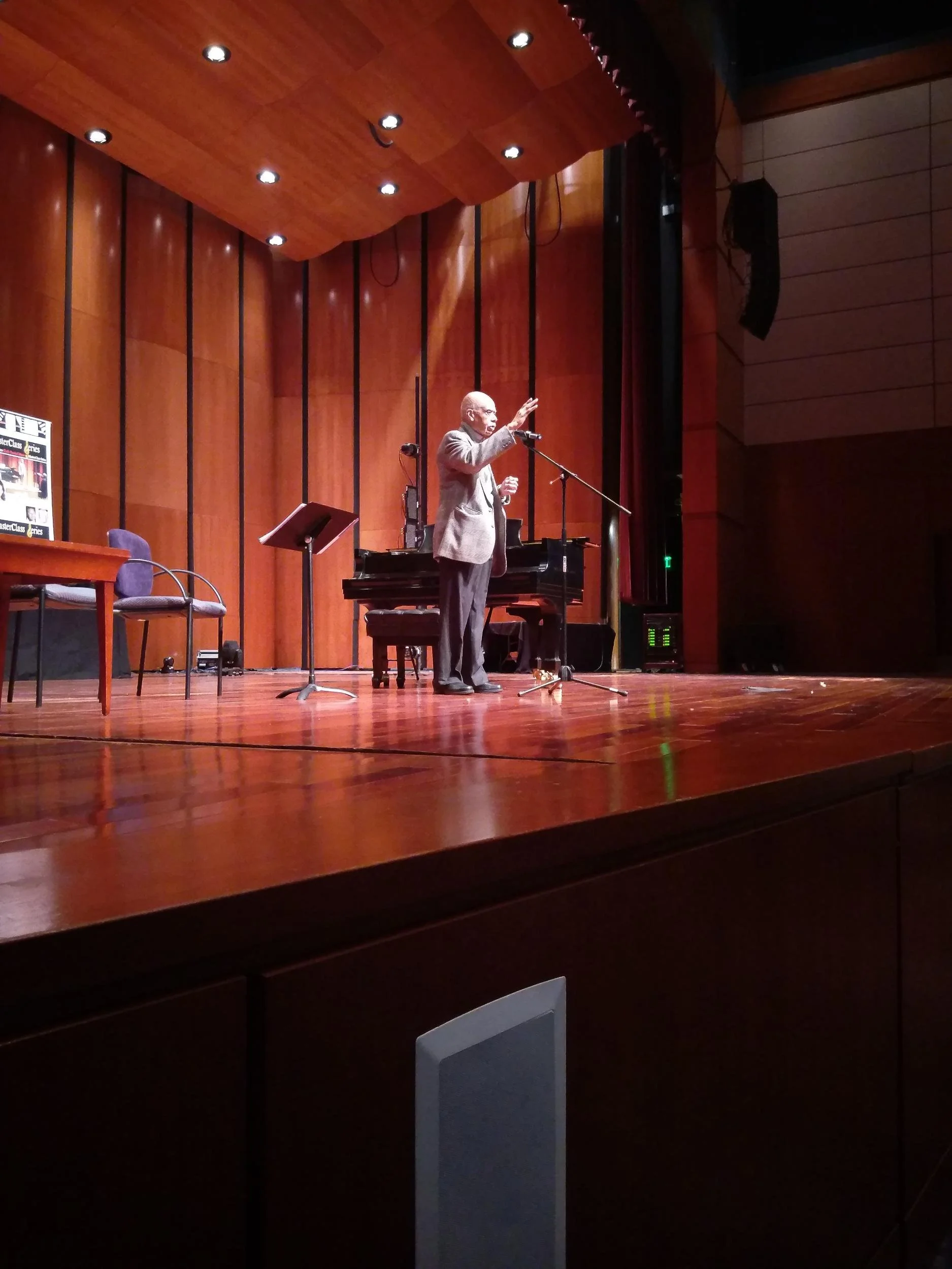 An elderly man stands on a wooden stage, speaking or performing into a microphone, with a grand piano behind him in a large auditorium with wooden panel walls.