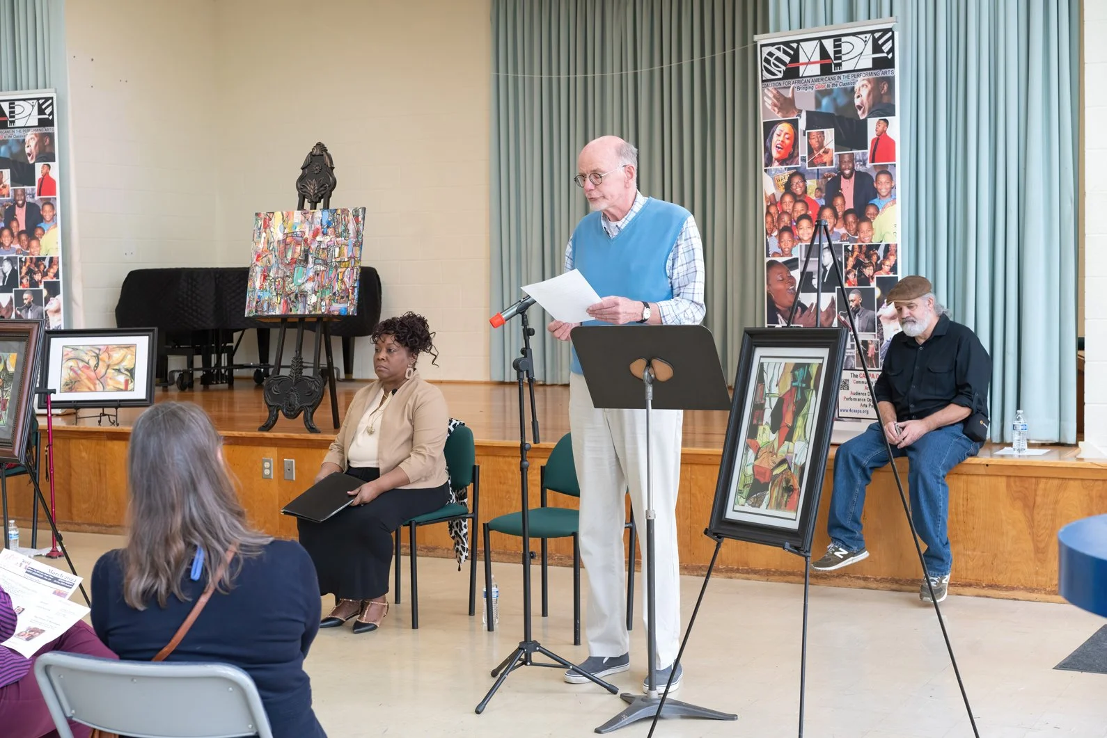 An elderly man with glasses and a blue vest speaking at a podium with a microphone during an art event, flanked by two seated individuals, with artwork displayed on easels and banners in the background.