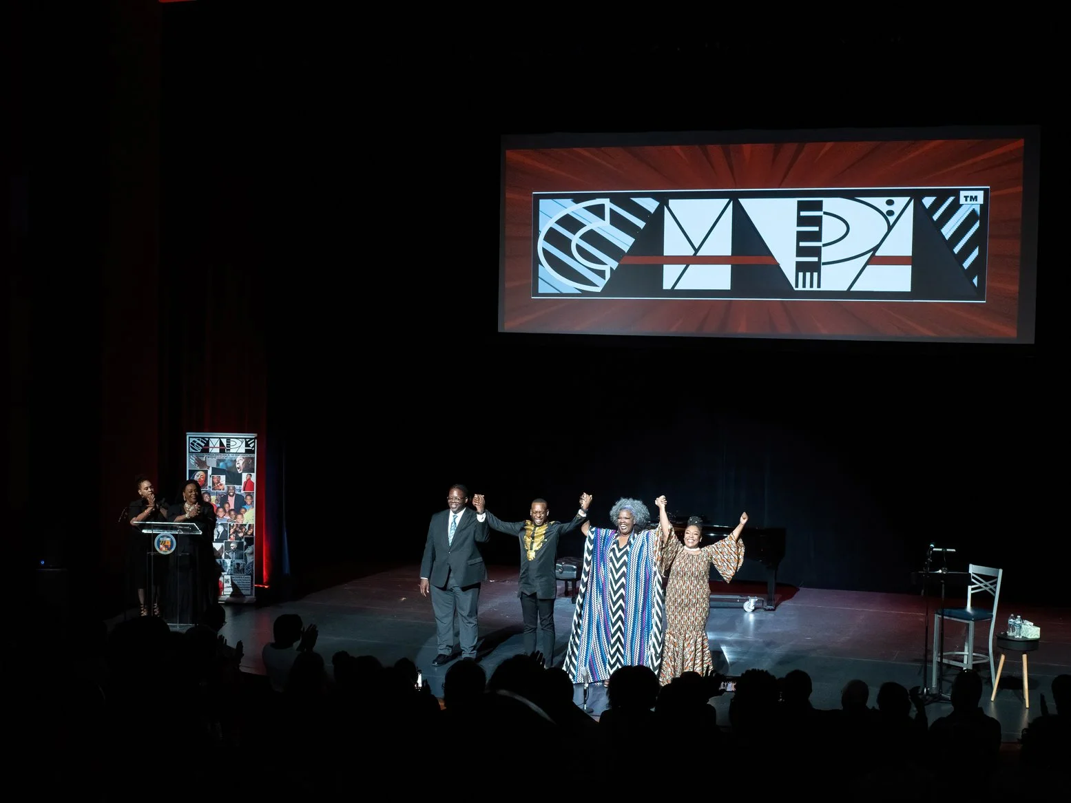 People on stage at a conference or event, with three individuals holding hands and celebrating, and a speaker at a podium to the left, with an audience in front.