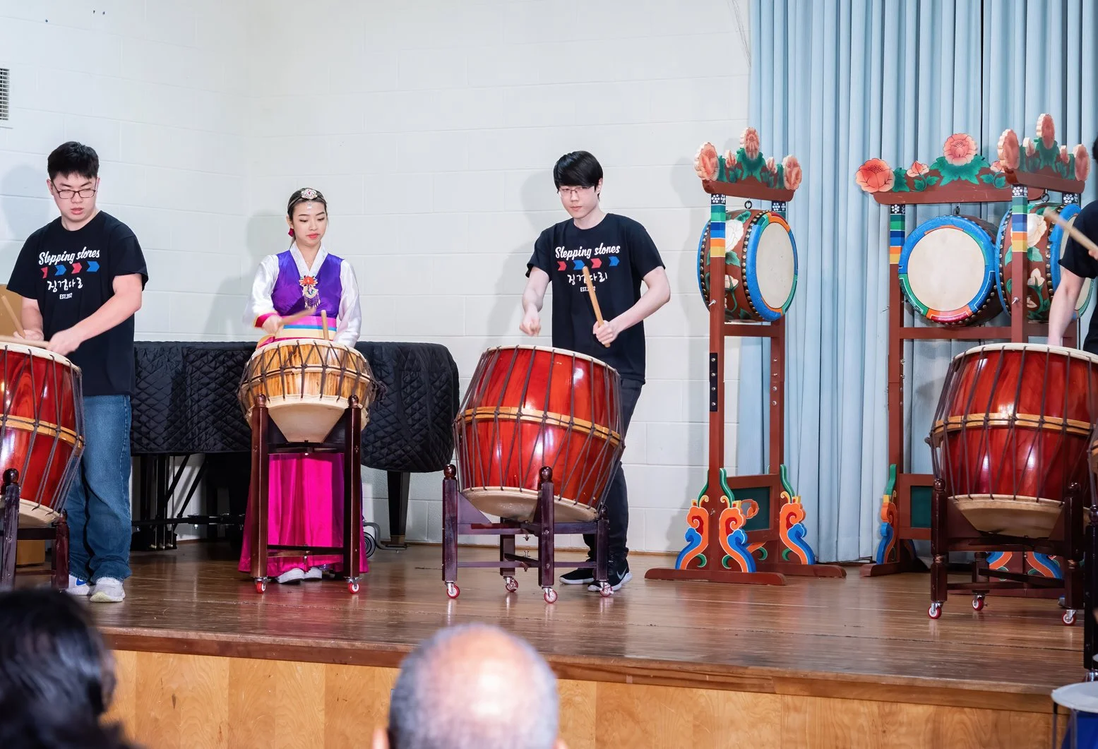 Group of people performing traditional Korean drum playing on stage with drums and decorative backdrop.